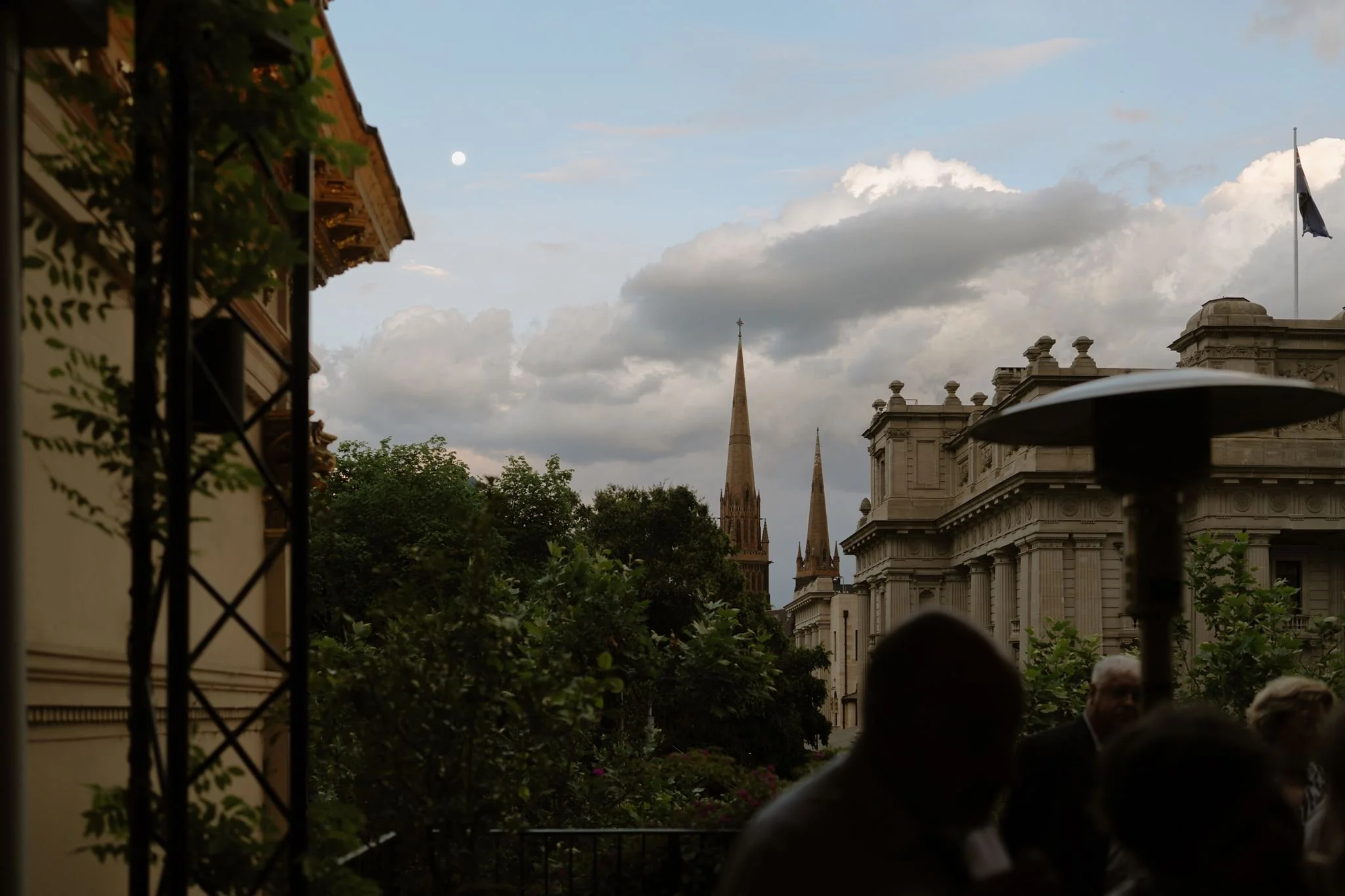 Cityscape view of historic buildings with spires and domes, partly obscured by trees, under a cloudy sky with a visible full moon.