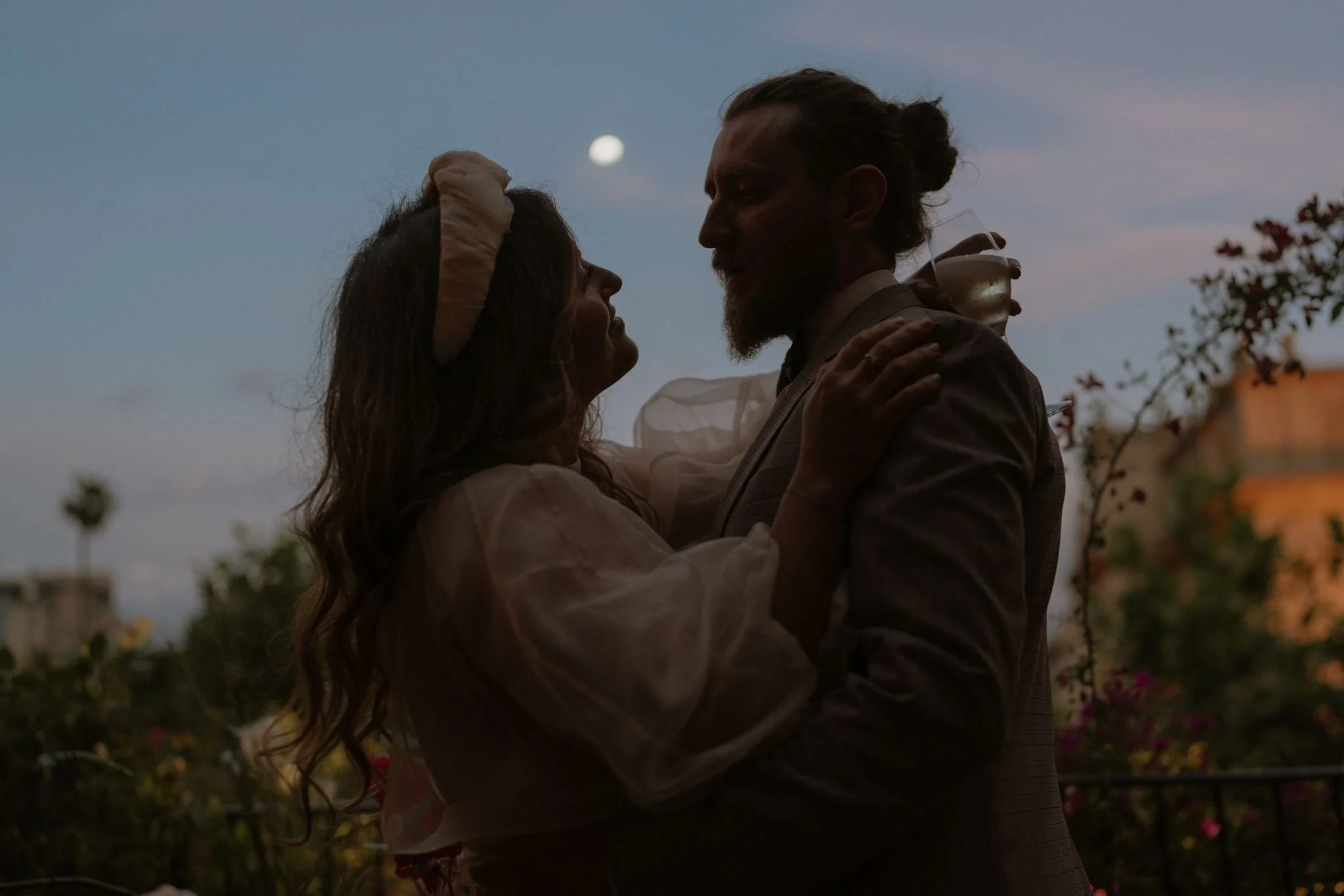 A man and woman embrace outdoor at dusk, with moon in the sky and trees and flowers in the background.