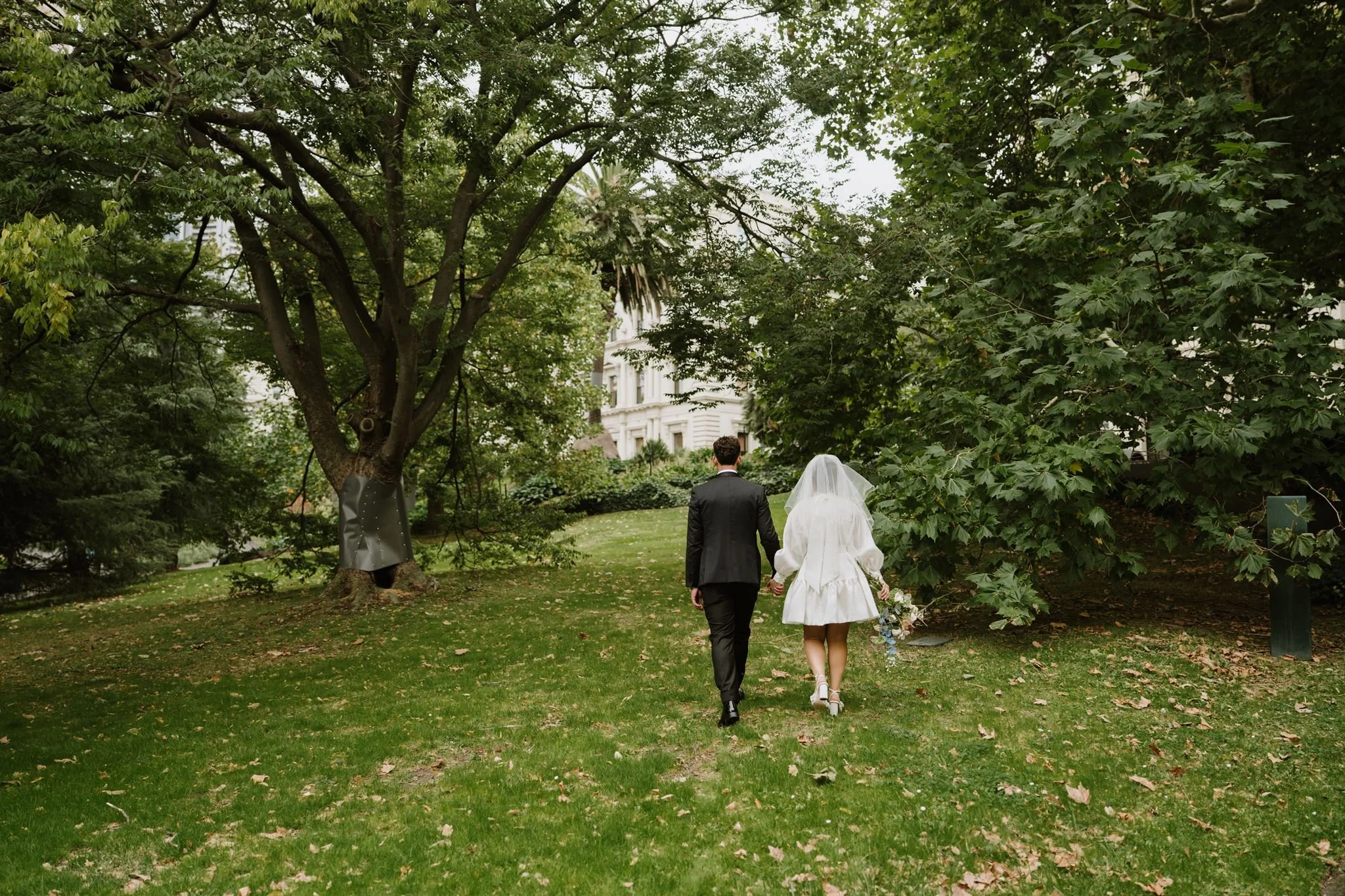 A bride and groom walking hand in hand through a park with trees and fallen leaves, with a large white building in the background.