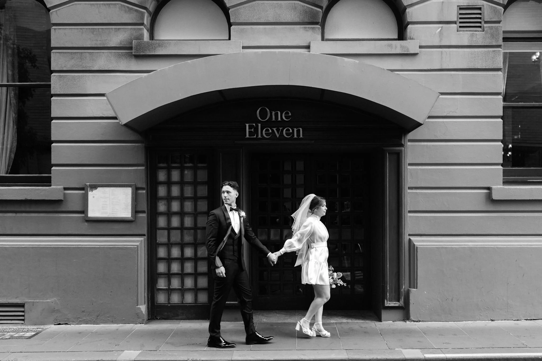 Black and white photo of a bride and groom holding hands while walking in front of a building with an arched entrance and the words 'One Eleven' above. The groom is in a tuxedo and the bride in a white dress and veil.