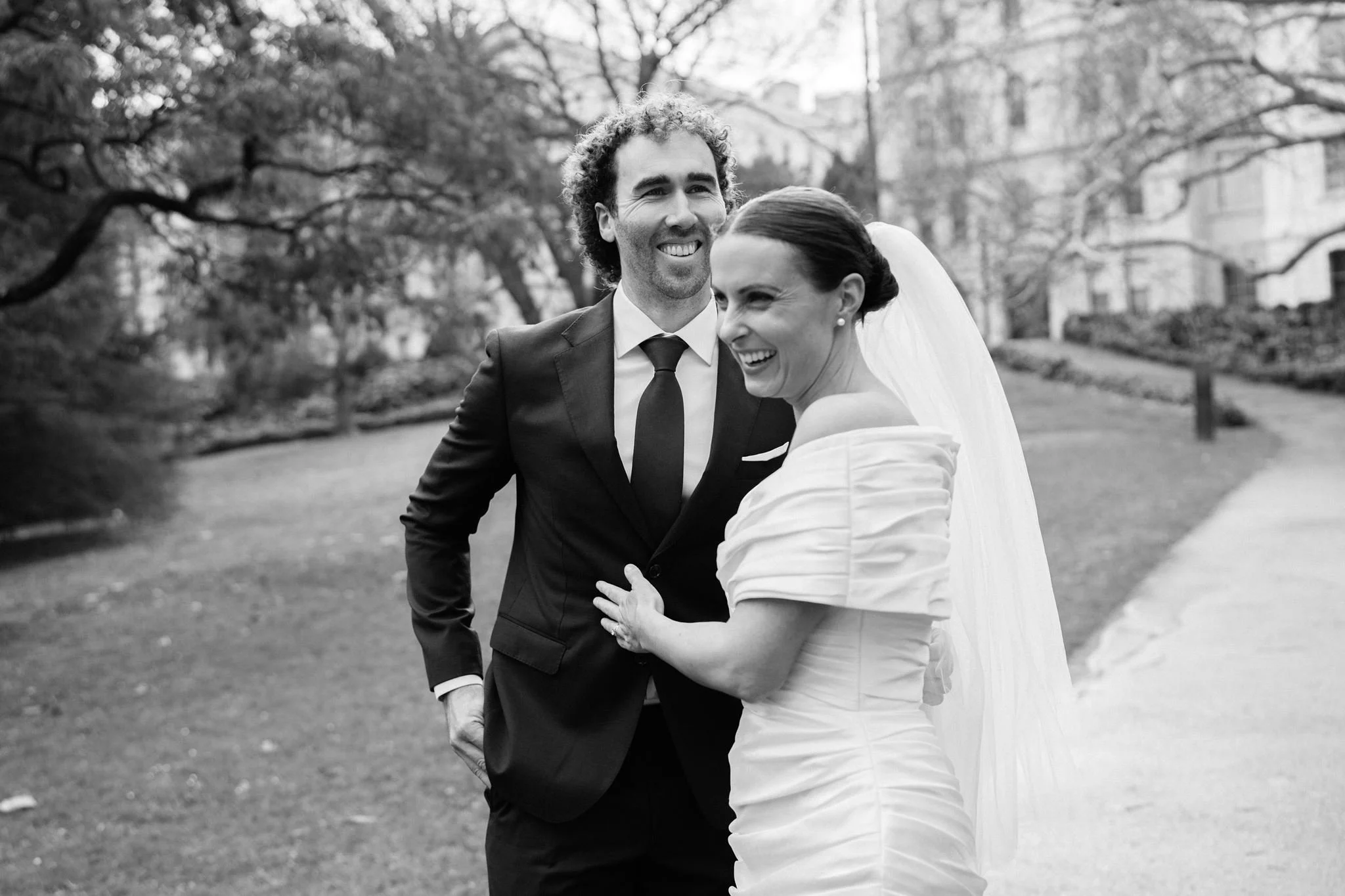 A black and white photo of a happy bride and groom outdoors, smiling and embracing each other.