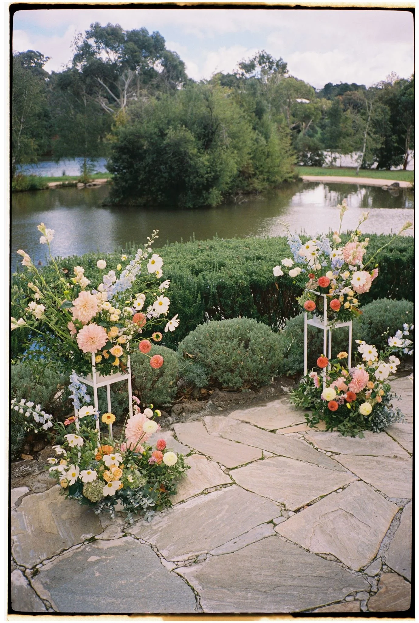 An outdoor scene with a pond, surrounded by green trees and a grassy area, with a pathway along the water. In the foreground, there are colorful flower arrangements on white stands and the stone-paved ground.