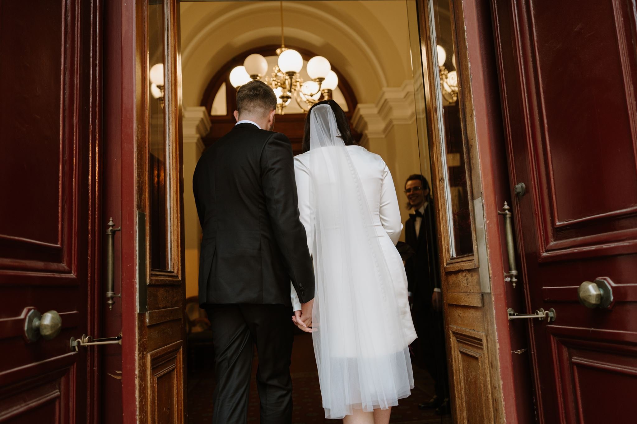 A bride and groom standing hand in hand inside a church, viewed from the back, with a priest in the background, seen through open red wooden doors, lit by ornate chandeliers.