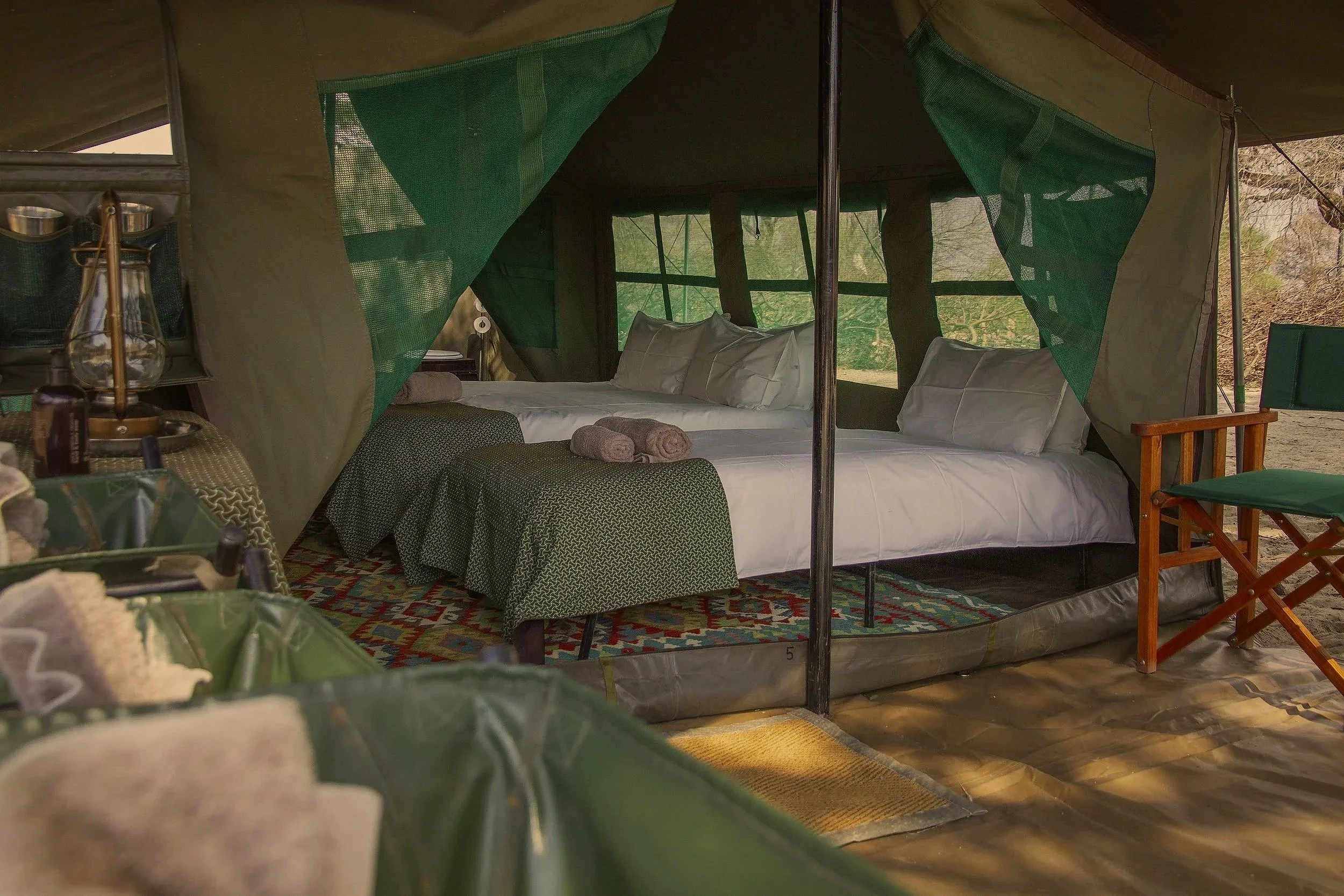 Inside a safari tent with two large beds covered with white linens and pillows, green patterned bed skirts, and rolled towels on top. The tent has mesh windows and green accents, with outdoor surroundings visible through the windows. There is a wooden chair and a small area with bottles and a lamp nearby.