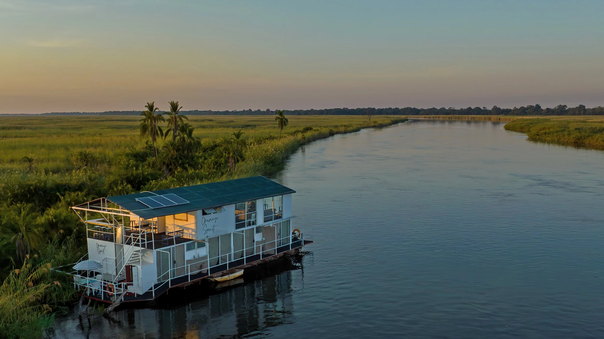 A houseboat on a river near lush green trees with a rainbow visible in the cloudy sky.