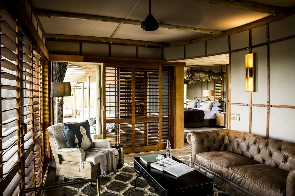 A cozy living room with wooden accents, cream and brown furniture, a leather sofa, an upholstered armchair with pillows, a coffee table with a water bottle and open magazine, and a view into a bedroom with a bed and draped curtains.
