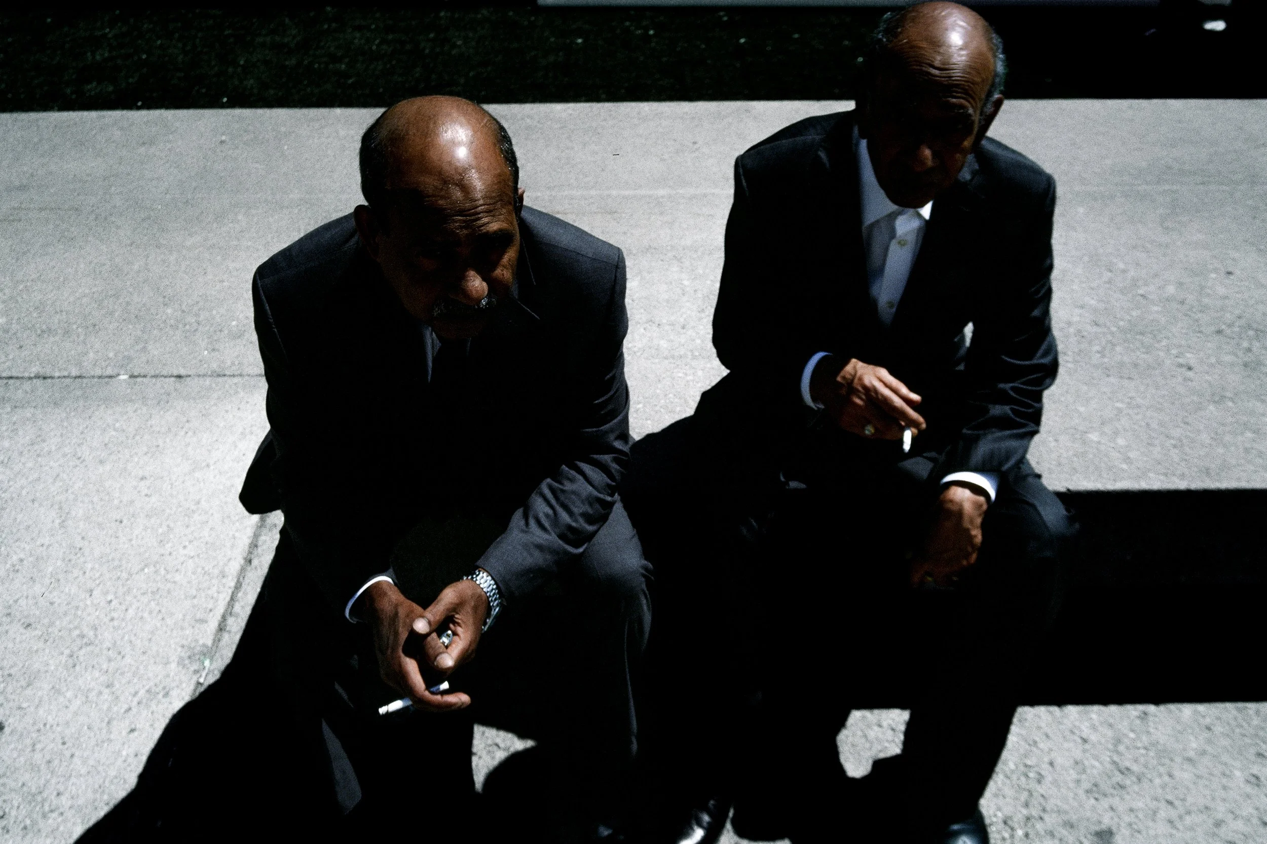 Two elderly men wearing dark suits are squatting on a concrete sidewalk at night. One man is holding a cigarette in his right hand and both are engaged in a serious conversation.