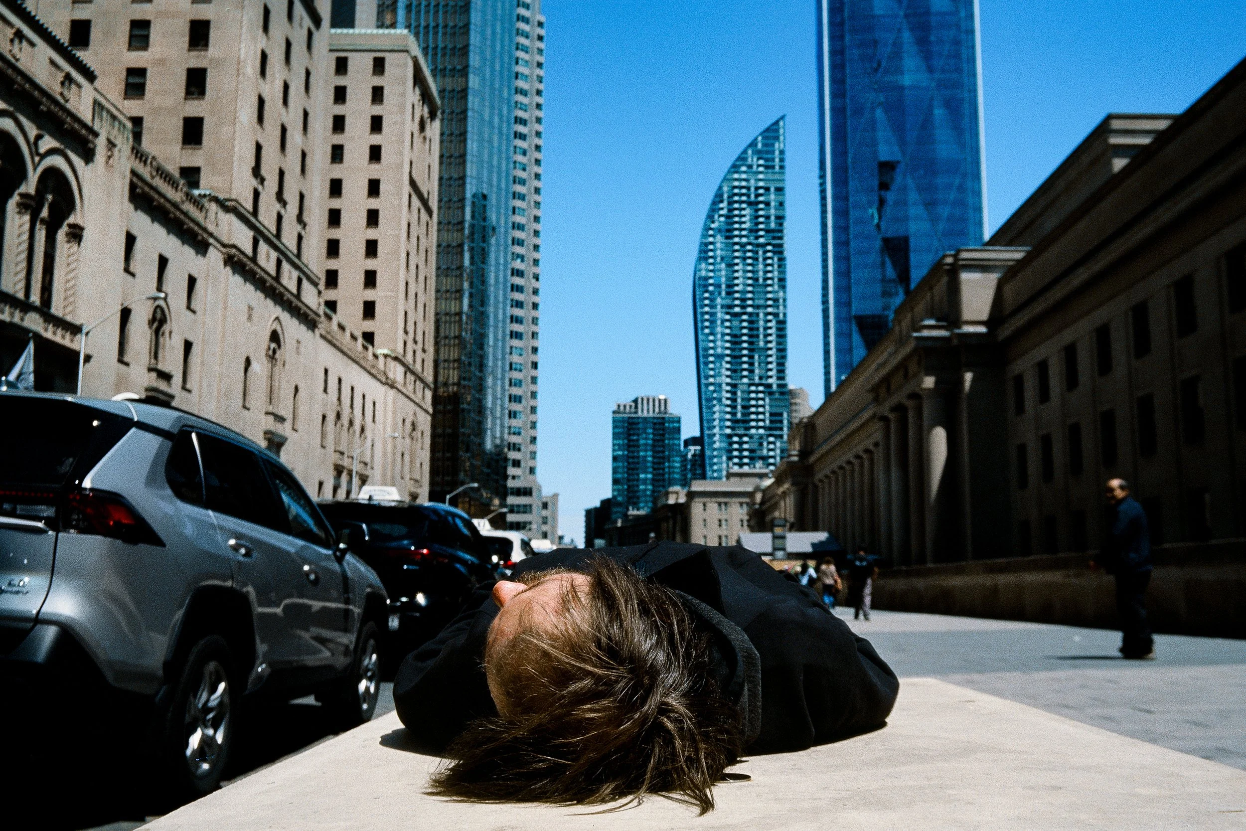 A person has fallen on the ground in an urban street with tall buildings, cars parked along the street, and a clear blue sky.
