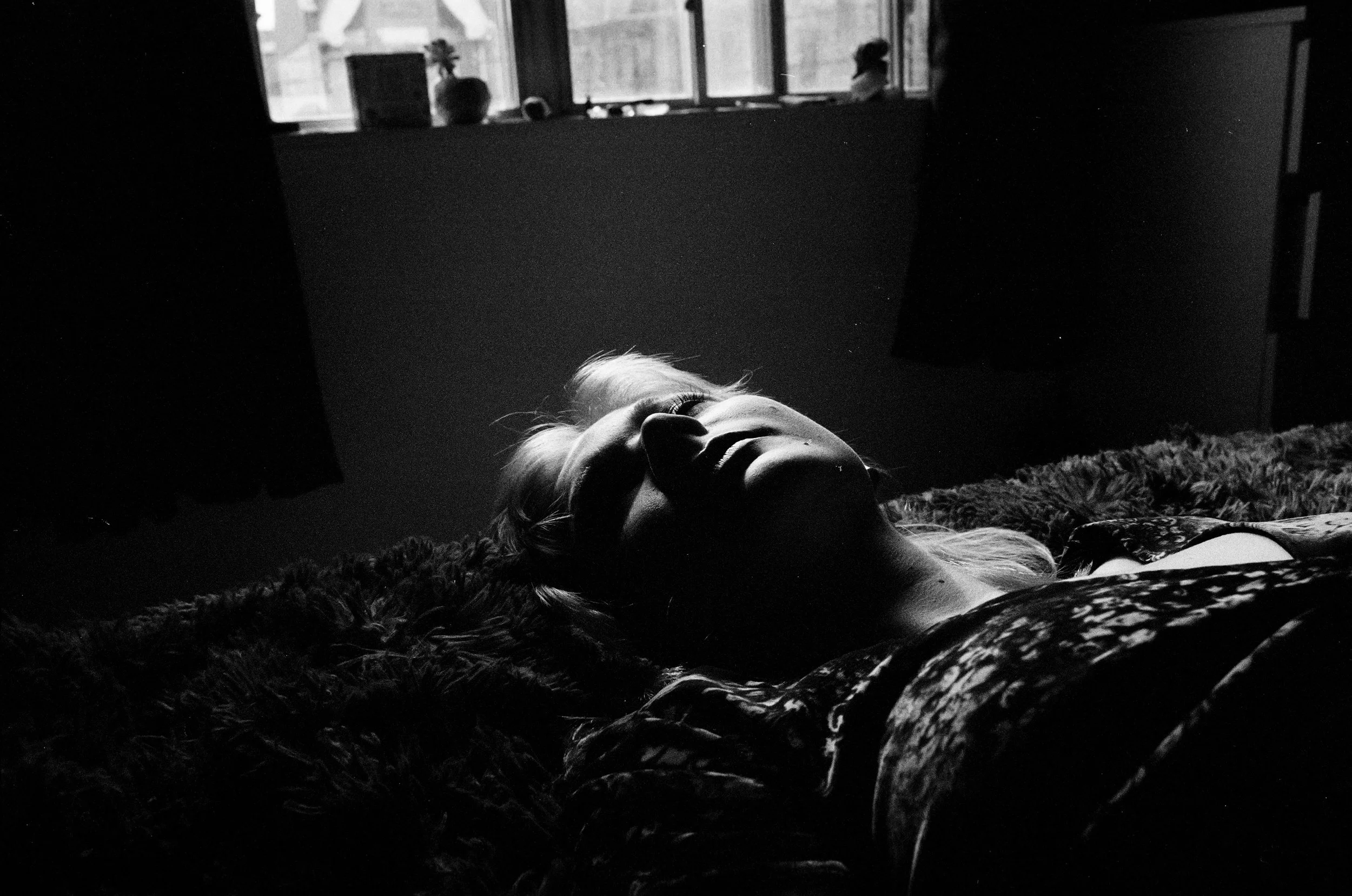 Black and white photo of a woman lying on her back on a bed, with her face illuminated by light from a window behind her.