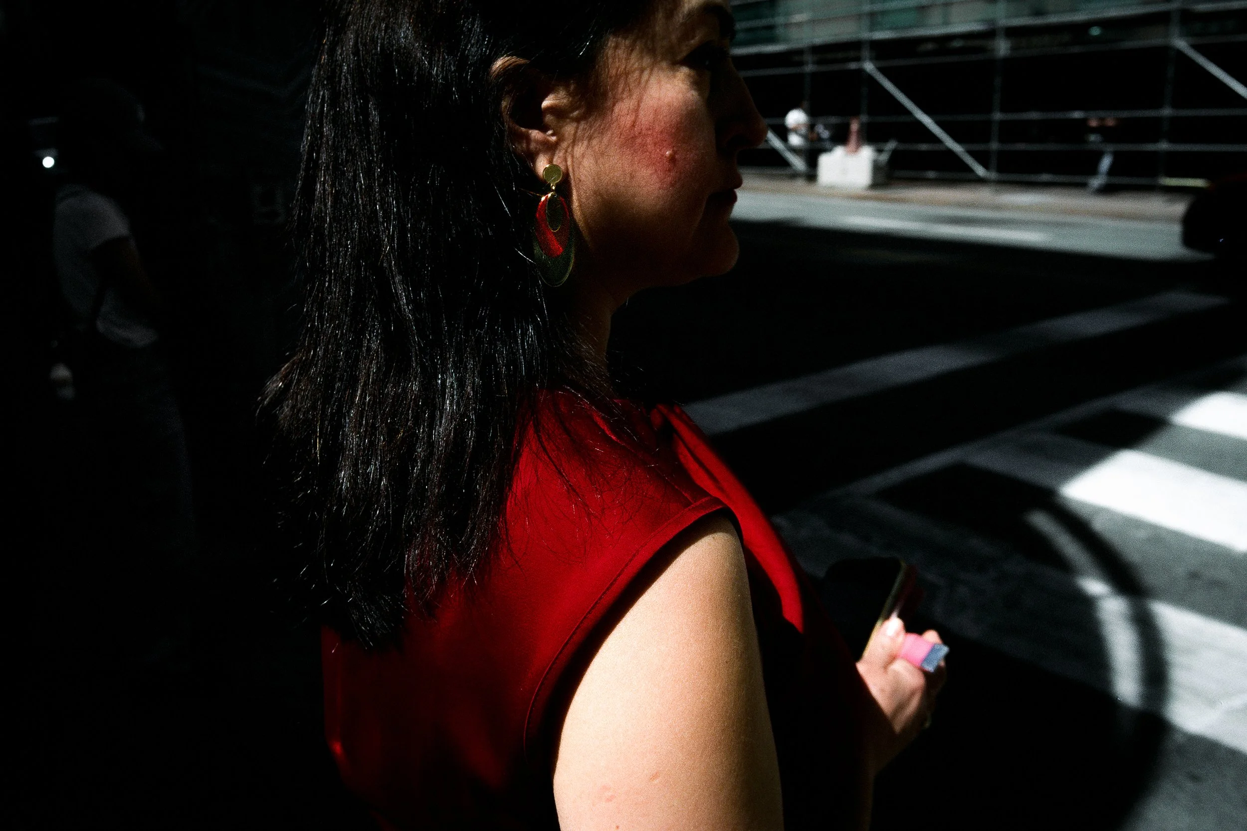 Profile of a woman with black hair wearing large colorful earrings and a red top, standing near a crosswalk at night.