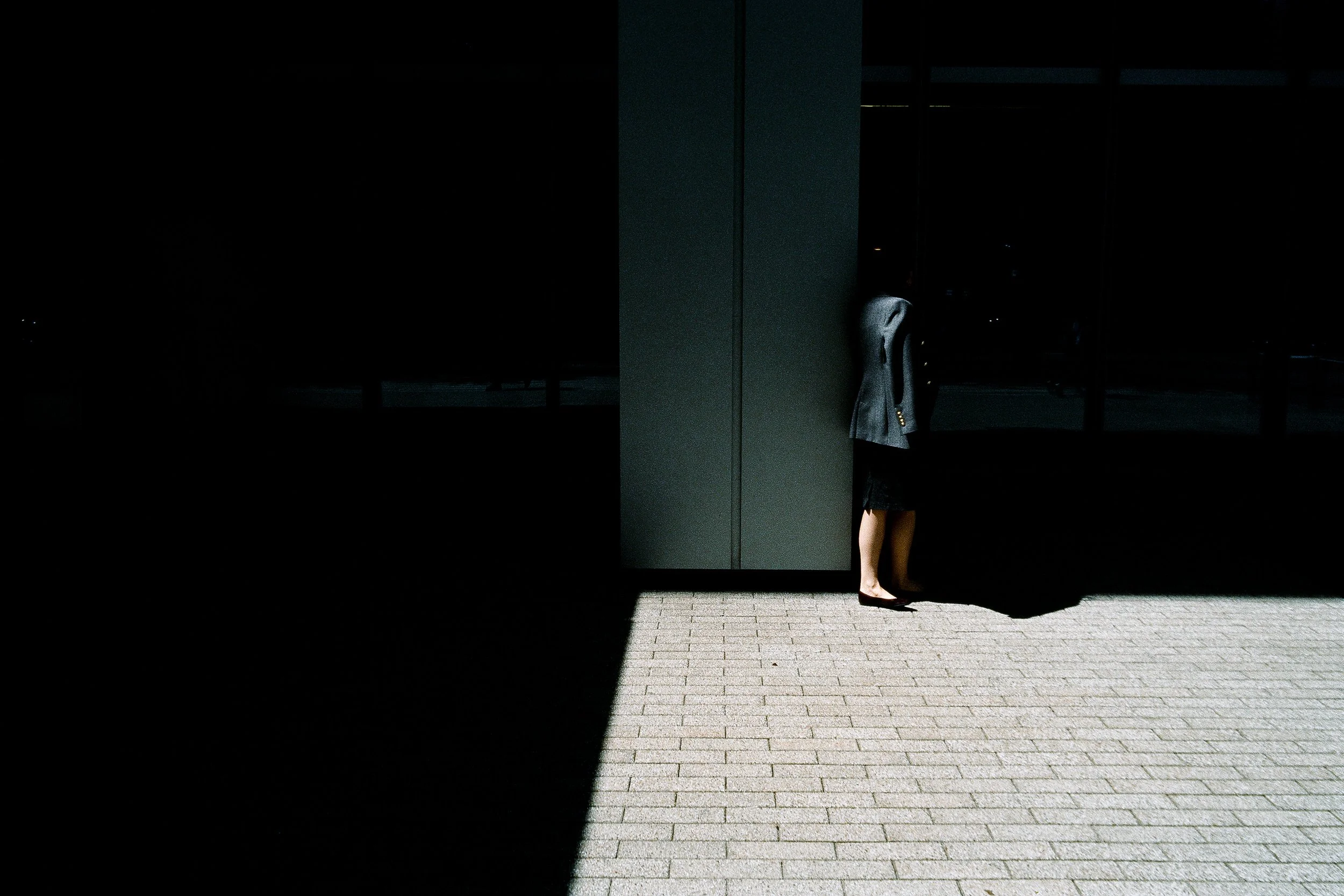 A woman in a dark blazer and skirt standing on a sunny sidewalk, partially concealed behind a large dark column outside a building with glass windows at night.
