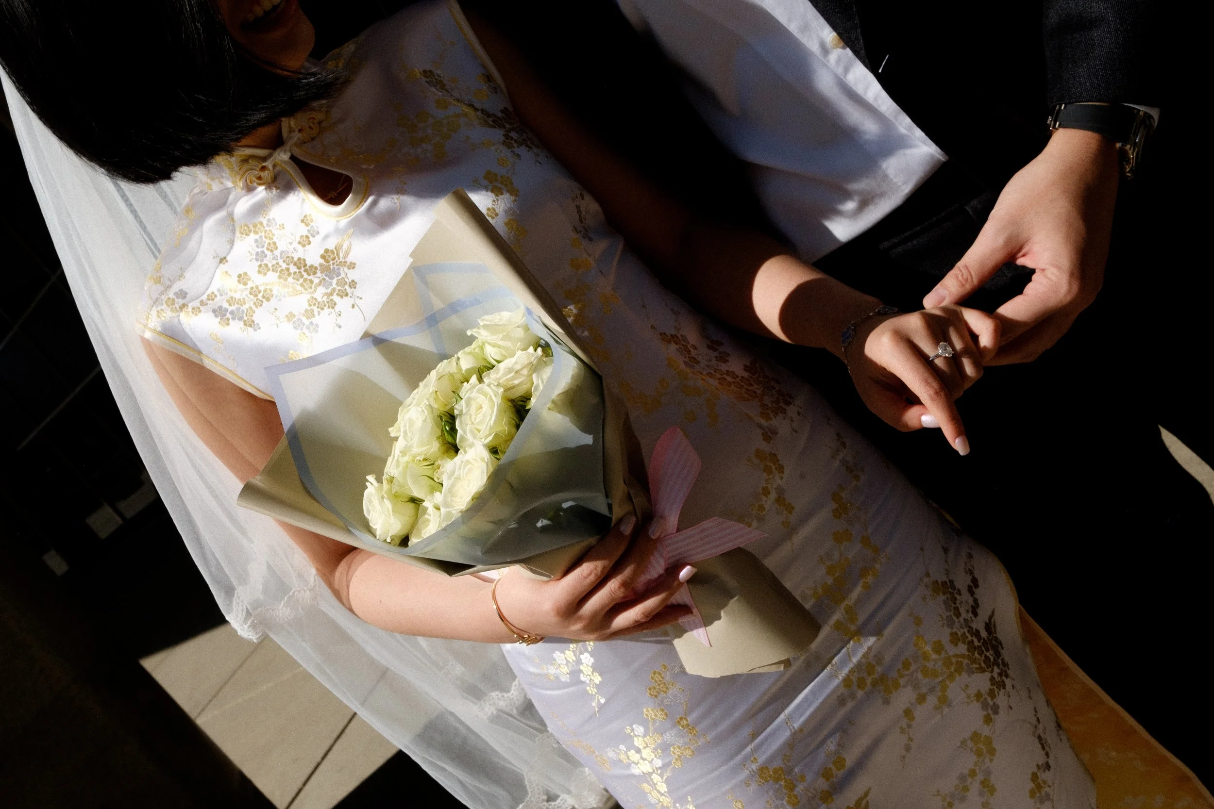 A person wearing a wedding dress holding a bouquet of white roses, standing at a table covered with a decorative cloth, with only their upper body and hands visible.