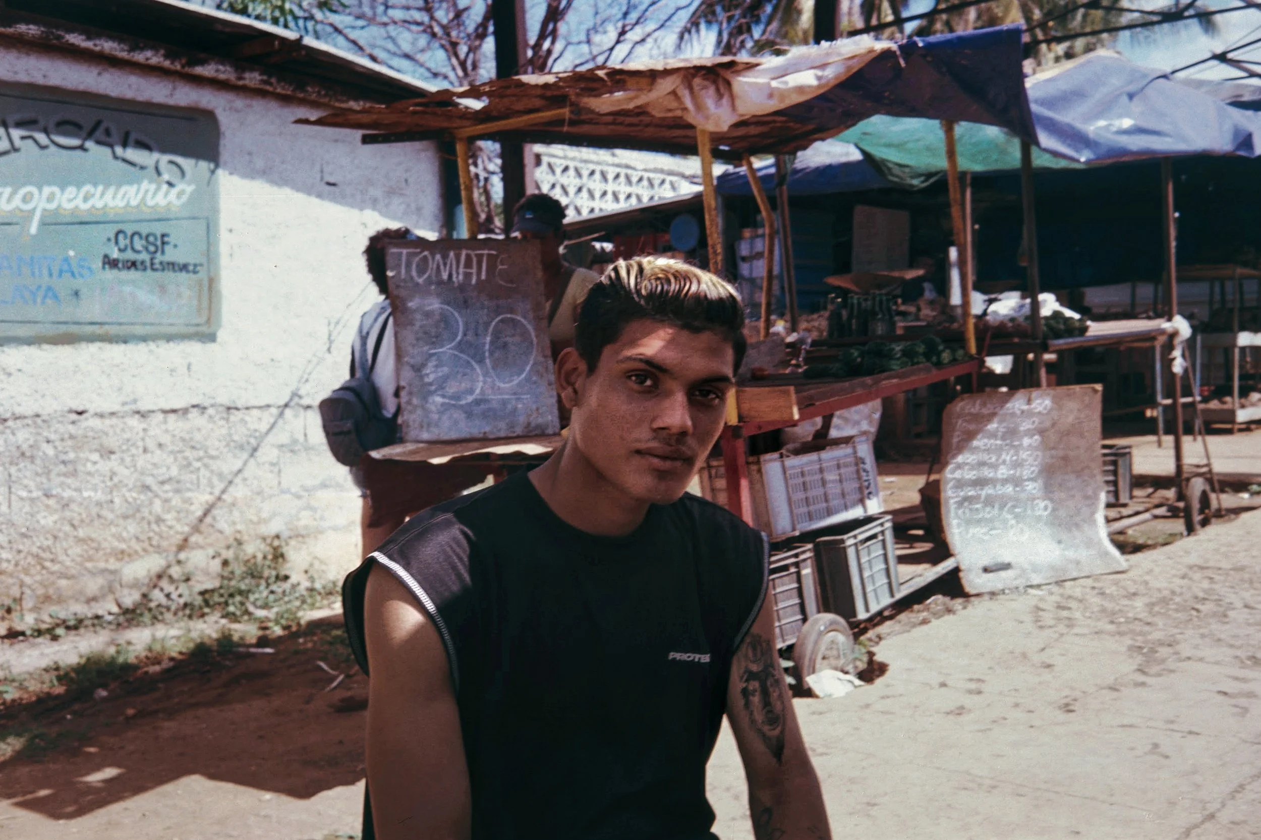 Young man with short dark hair and a tattoo on his left arm standing in front of a roadside stall selling tomatoes, with a sign indicating priced at 30. The stall is covered with a makeshift roof, and other items are visible on the shelves. A man in 