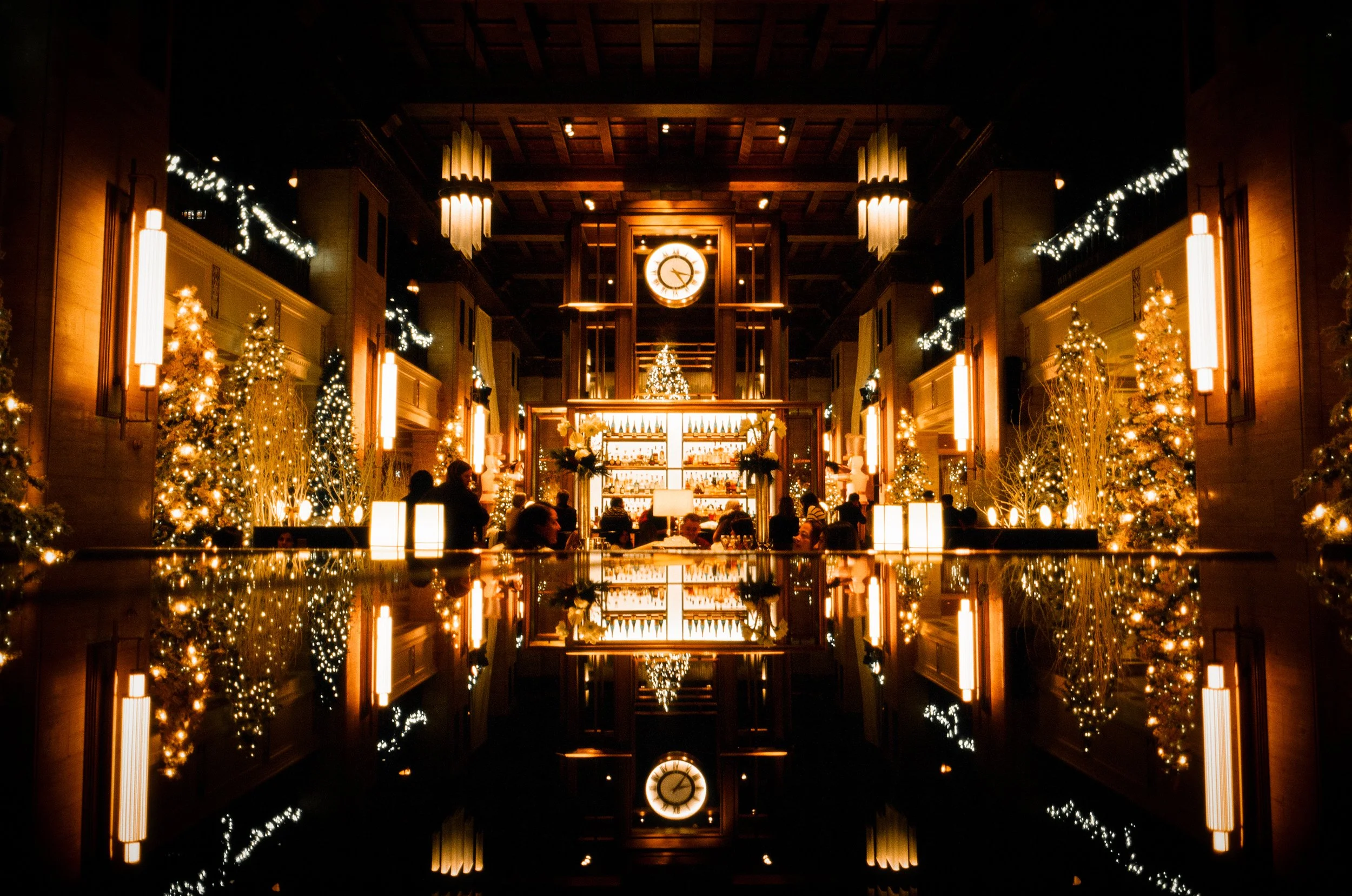 A warmly lit, festive indoor space with Christmas trees decorated with lights and ornaments, a large clock on the wall, and a bar in the background, reflecting on a shiny surface below.
