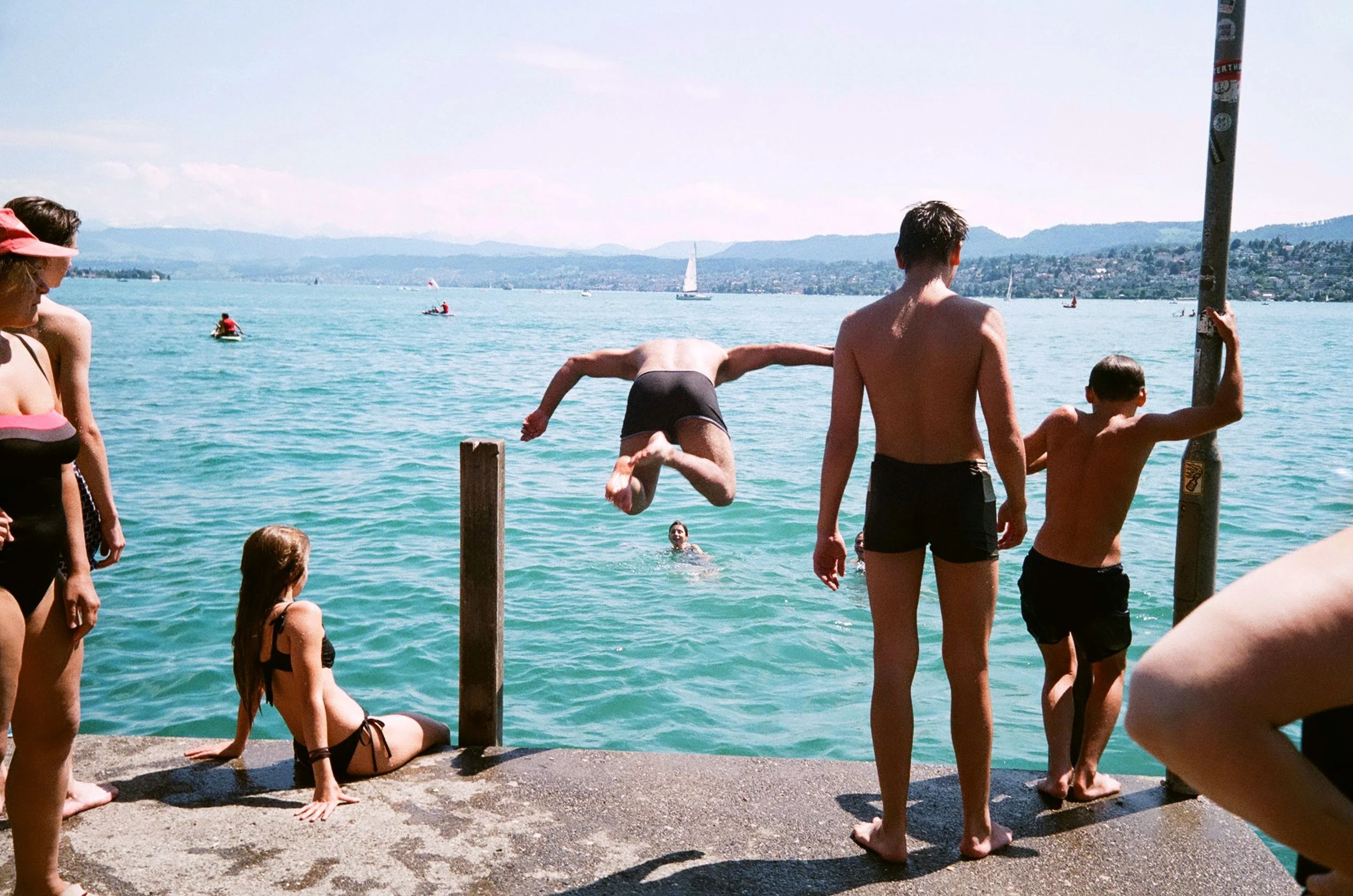 People at a lakeside dock with some jumping into the water and others waiting to swim, facing the lake with sailboats and a hilly landscape in the background.