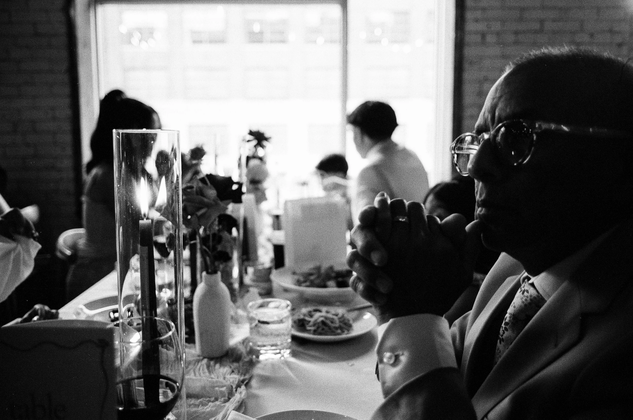 A woman with glasses and earrings is sitting at a table with a glass of red wine, holding her hand in a prayer position near her face. Several people can be seen sitting at the table in the background, and the setting appears to be a restaurant or ca