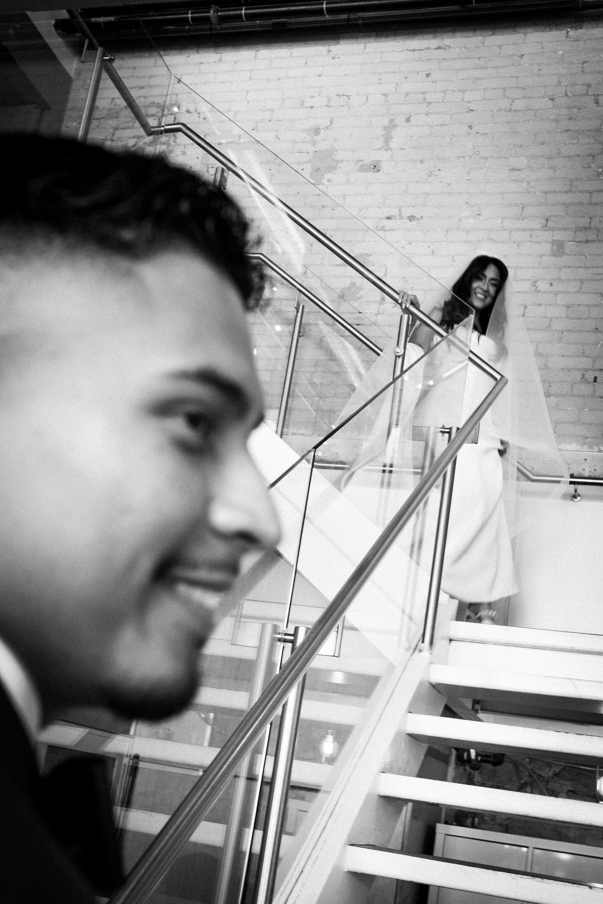 A black and white photo captures a smiling woman in this image, with a man's profile in the foreground and stairs with glass railings in the background.