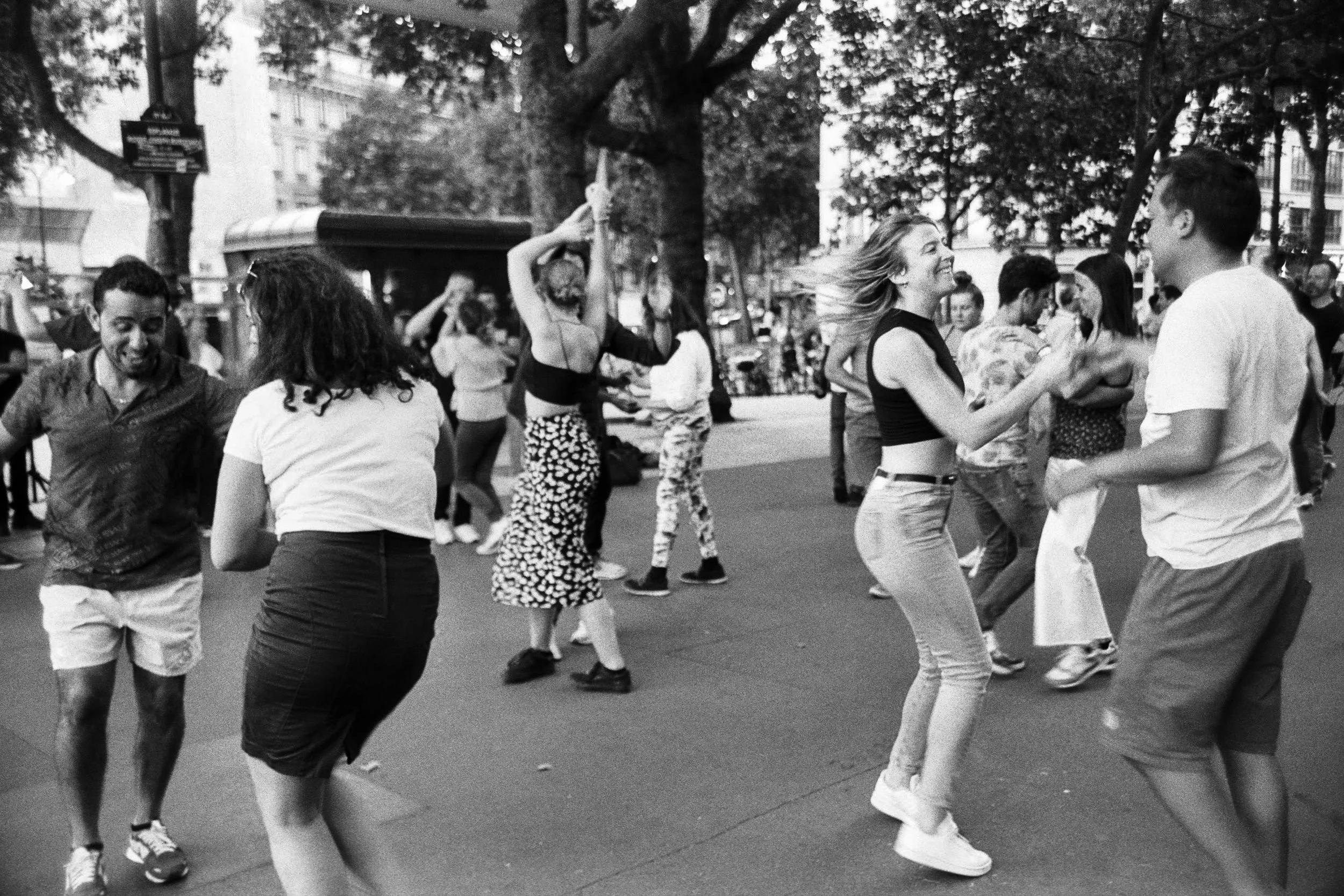 A group of people dancing outdoors on a city street surrounded by trees and buildings. The photo is in black and white.