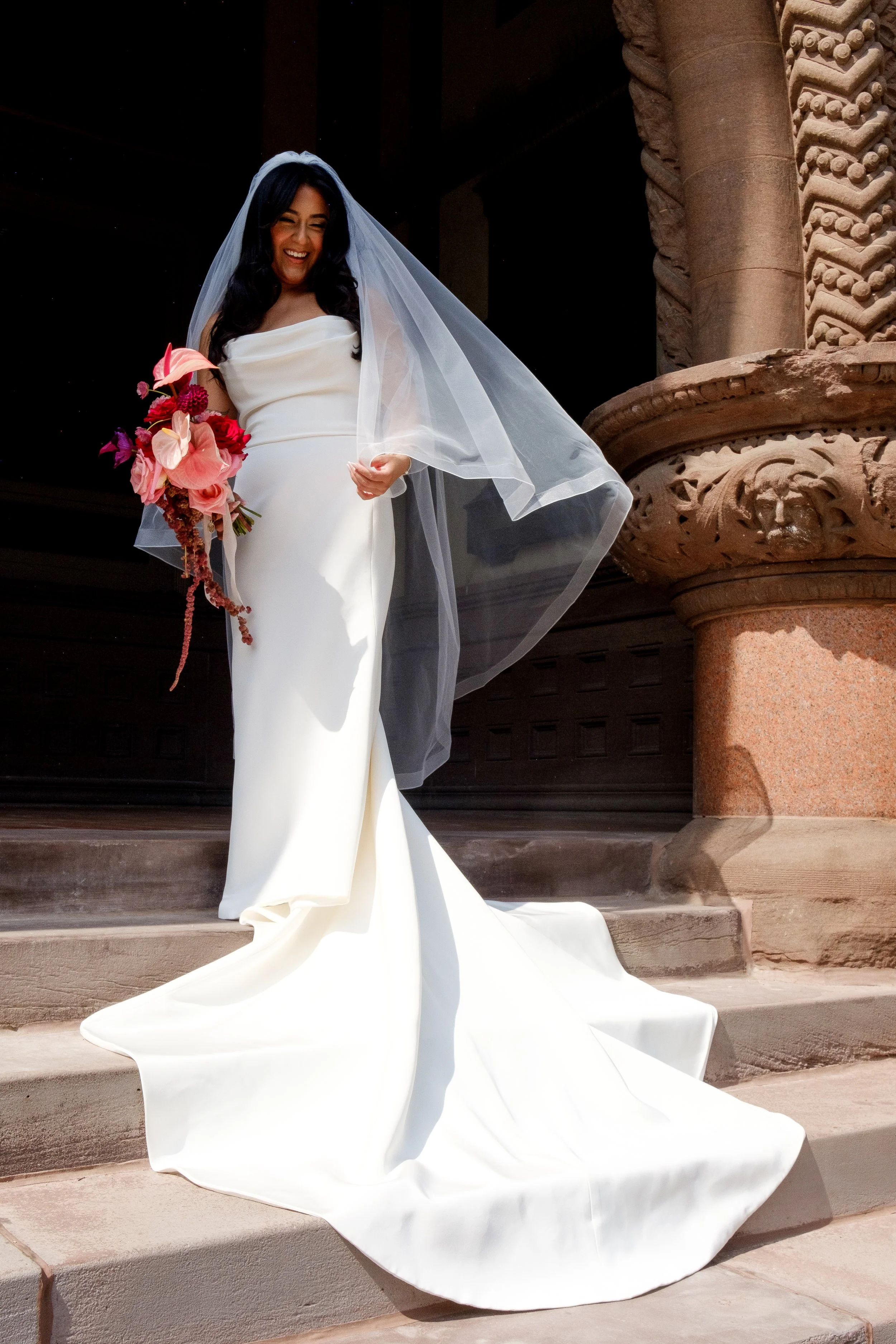 A smiling woman in a white wedding dress with a long train and veil, holding a bouquet of pink and purple flowers, standing on steps outside a stone building with intricate carvings.