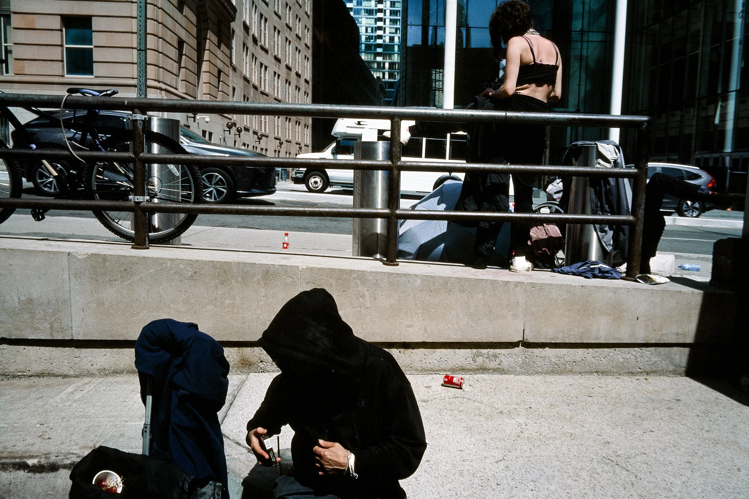 People sitting on steps near a city street with parked cars and tall buildings in the background.