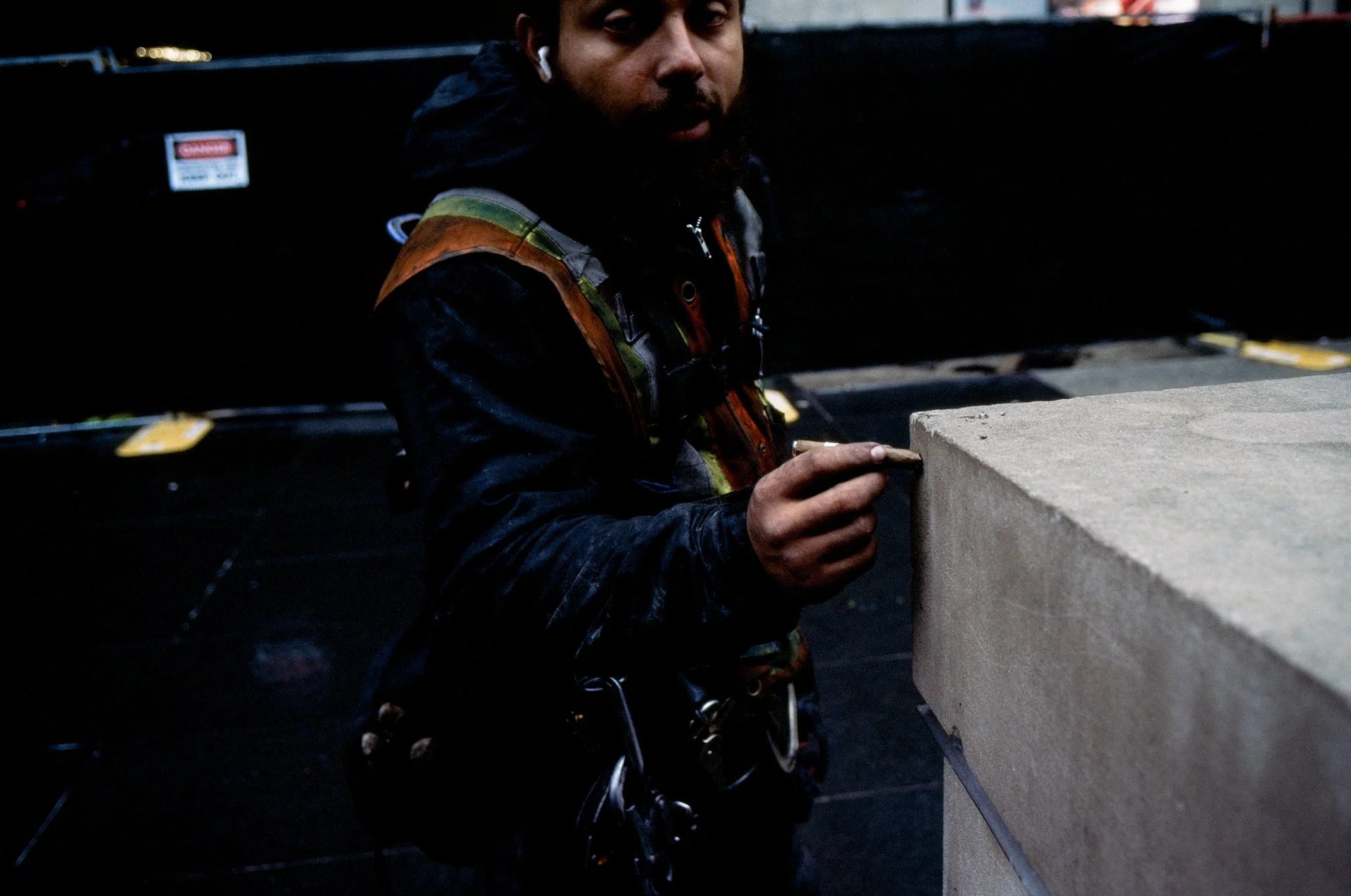 A man wearing safety gear and a harness is working on a construction project, using a tool on a concrete surface in an indoor setting.