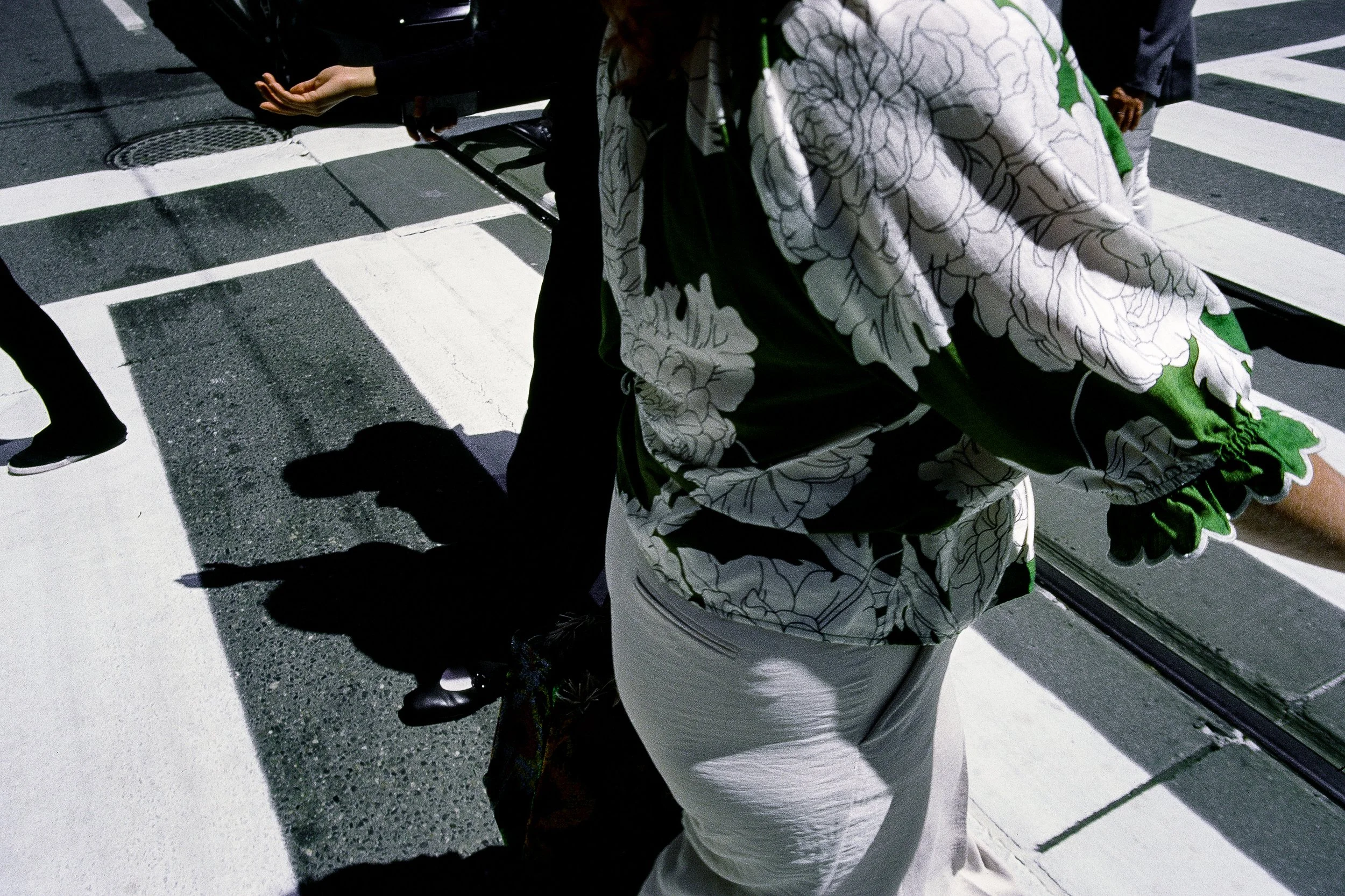 People walking across a street at night with white crosswalk lines, some of the individuals are dressed in dark clothing, and one person is wearing a floral patterned jacket with white pants.