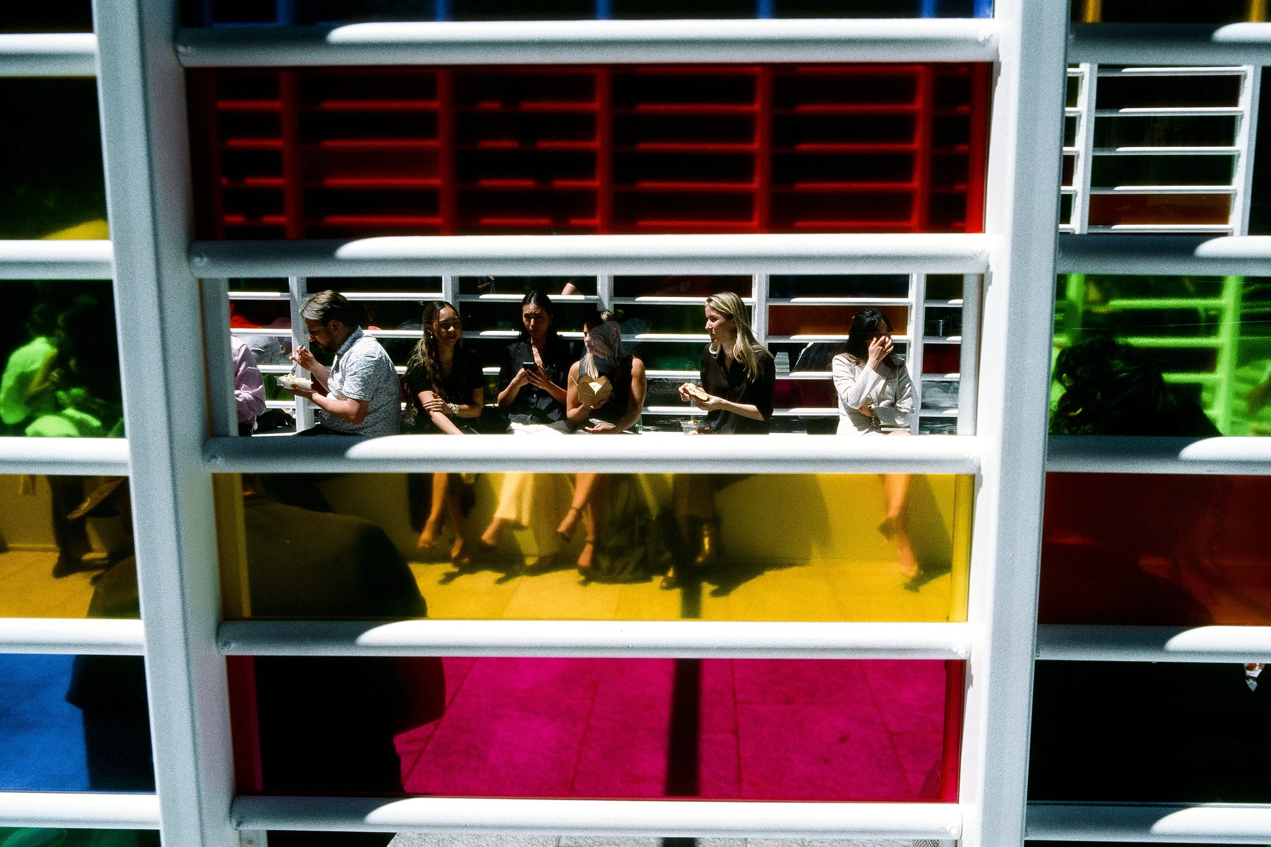 People sitting on a bench, through colorful window panes with red, yellow, green, and blue sections.
