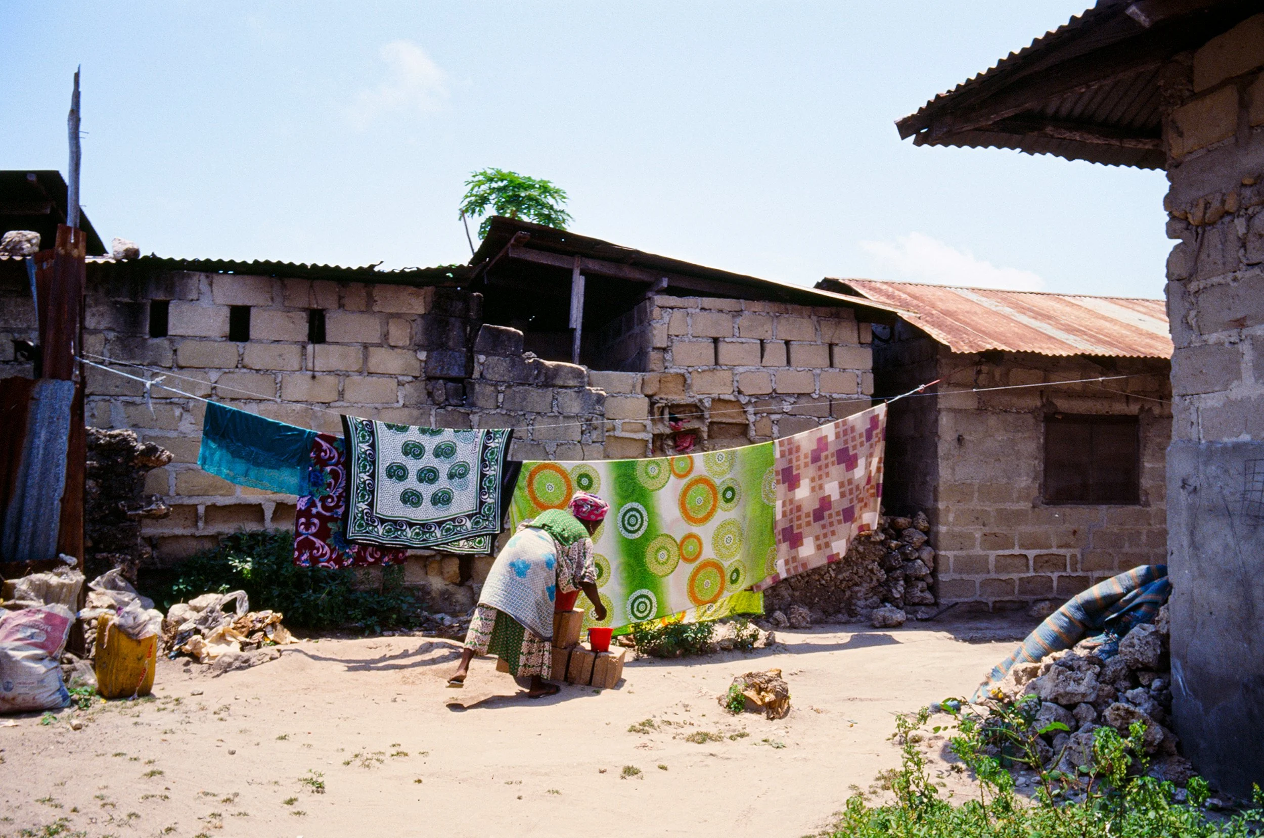 A woman hanging colorful laundry on a clothesline outside a rustic brick house with a corrugated metal roof.