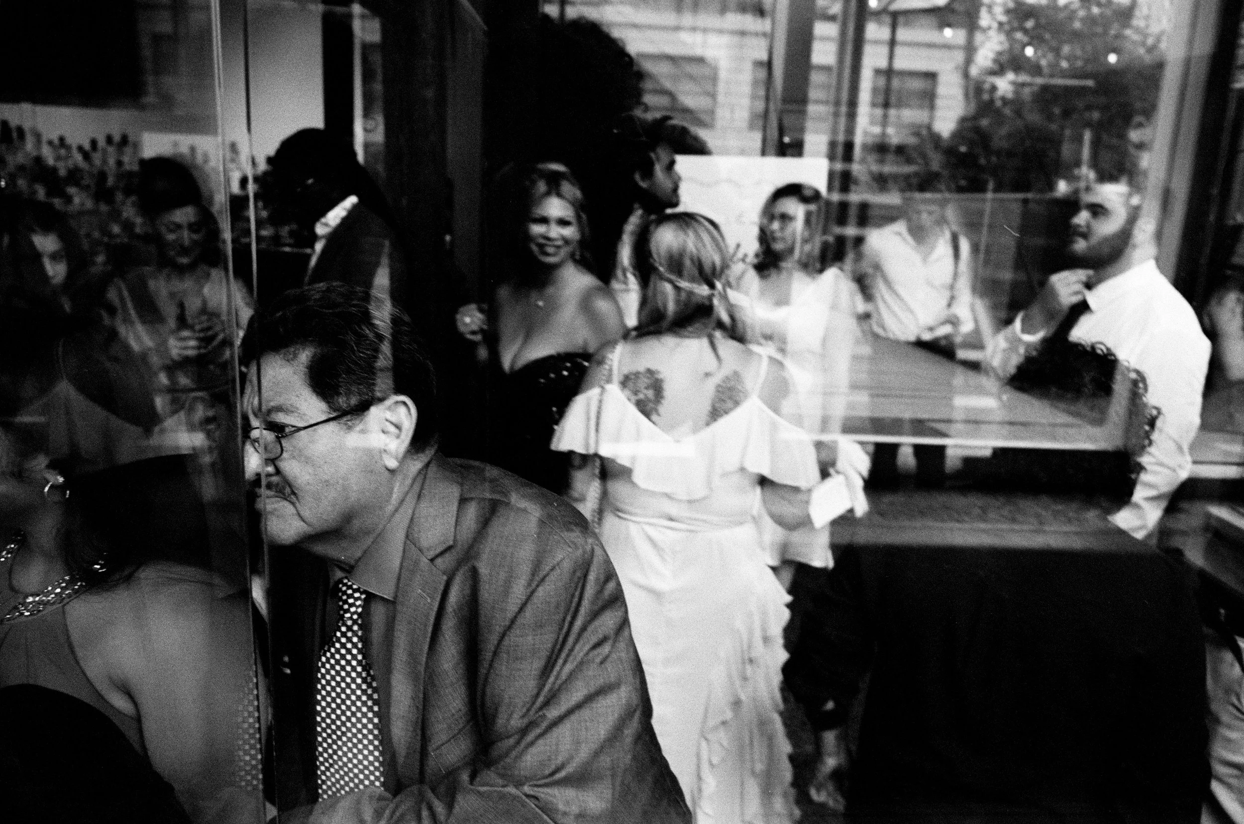 A group of people, mostly women, gathered in a room with one woman at a table in the foreground, and several others standing and talking behind her. The setting appears to be social or a meeting, seen through a glass reflection.