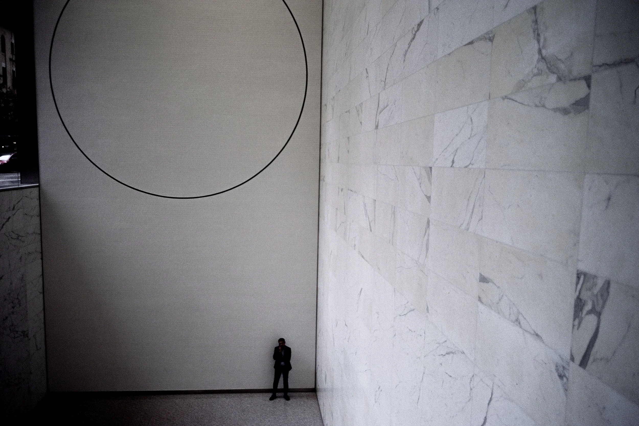 A person standing in a large, minimalist hall with white marble walls and floor, with a large circular outline on one wall.