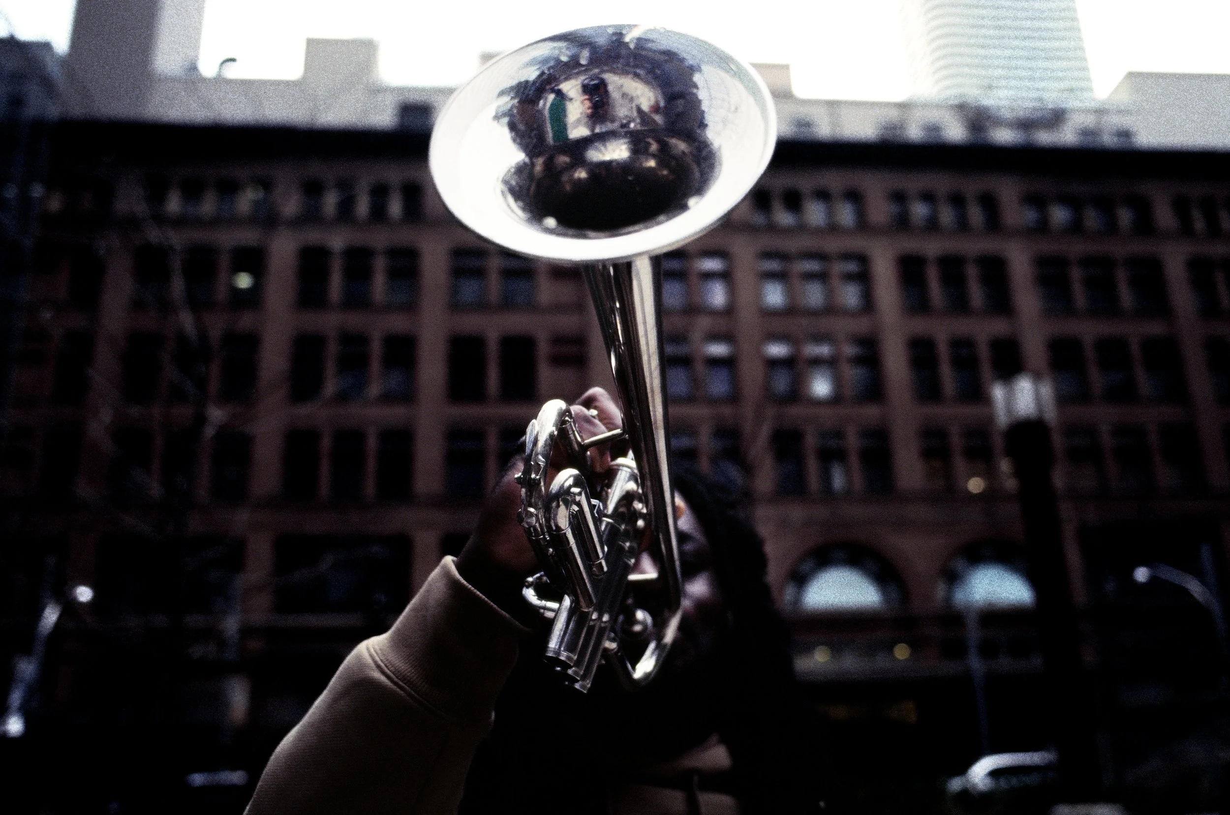 A hand holding a shiny silver trumpet against a cityscape background with tall buildings.