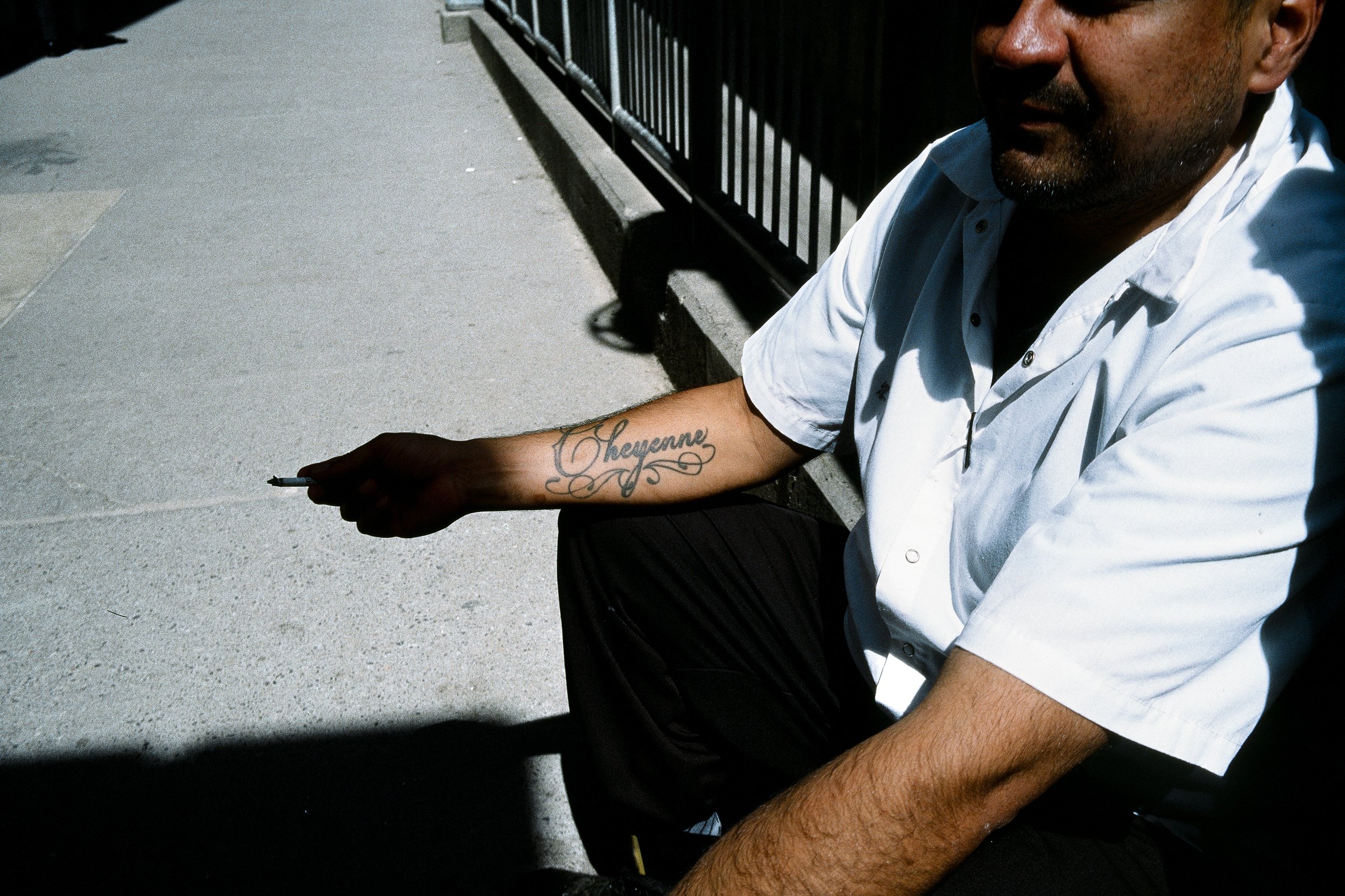 A man with a tattoo on his forearm sitting on the ground, holding a pen, wearing a white shirt, with a black fence in the background, partially visible on the right side of the image.