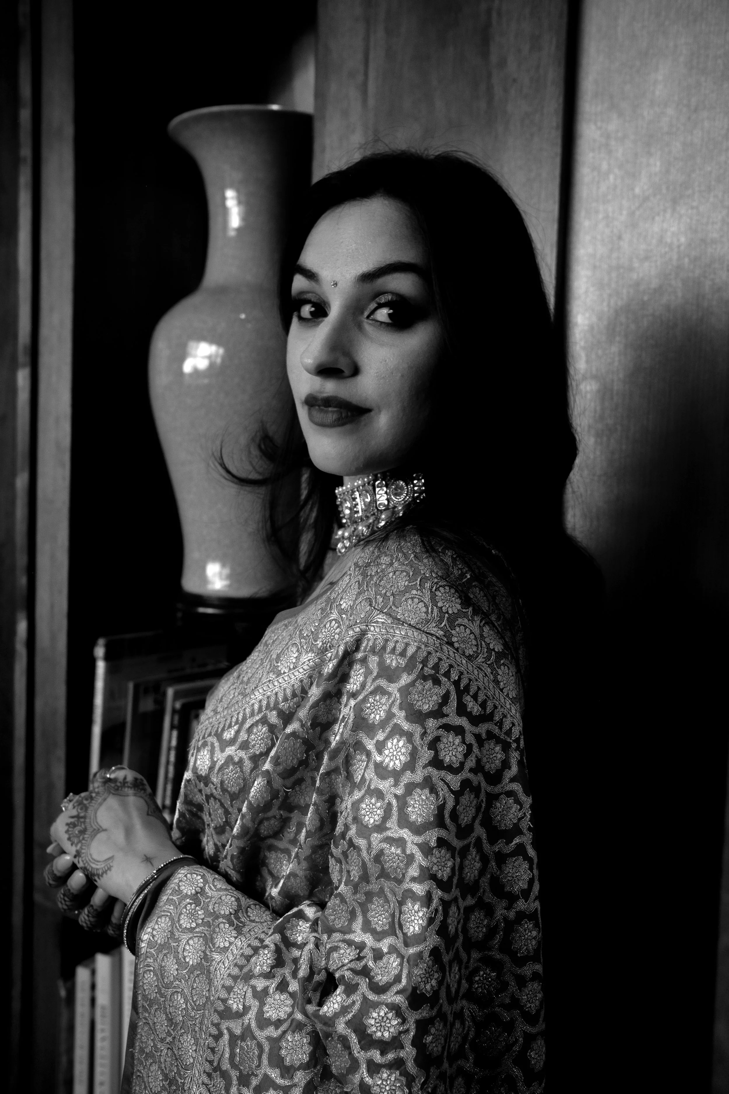 A woman wearing traditional Indian attire and jewelry, standing near a bookshelf and a large decorative vase, in black and white.