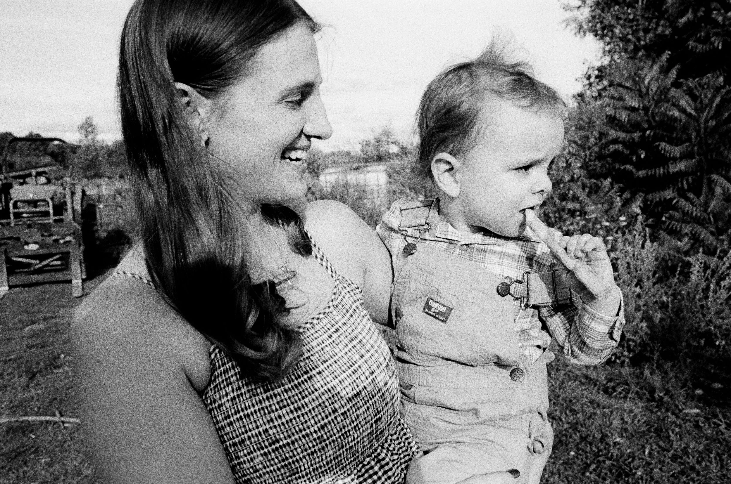 A woman and a young boy are outdoors, smiling and looking to the right, with trees and a fence in the background.