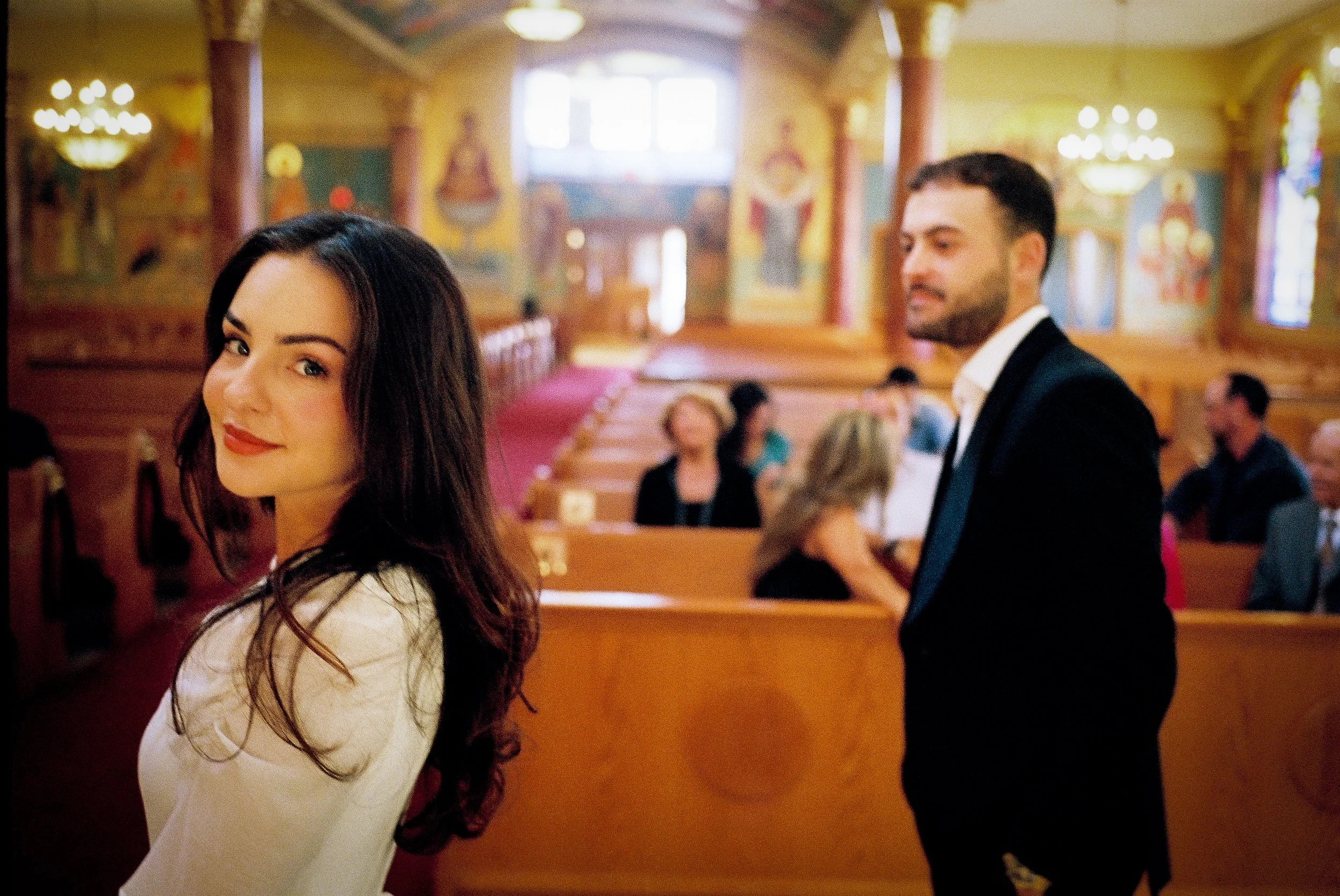 A woman with dark hair and a white top smiling and looking at the camera, with a man in a suit and white shirt standing nearby, inside a decorated church or hall with painted walls, chandeliers, and other people in the background.