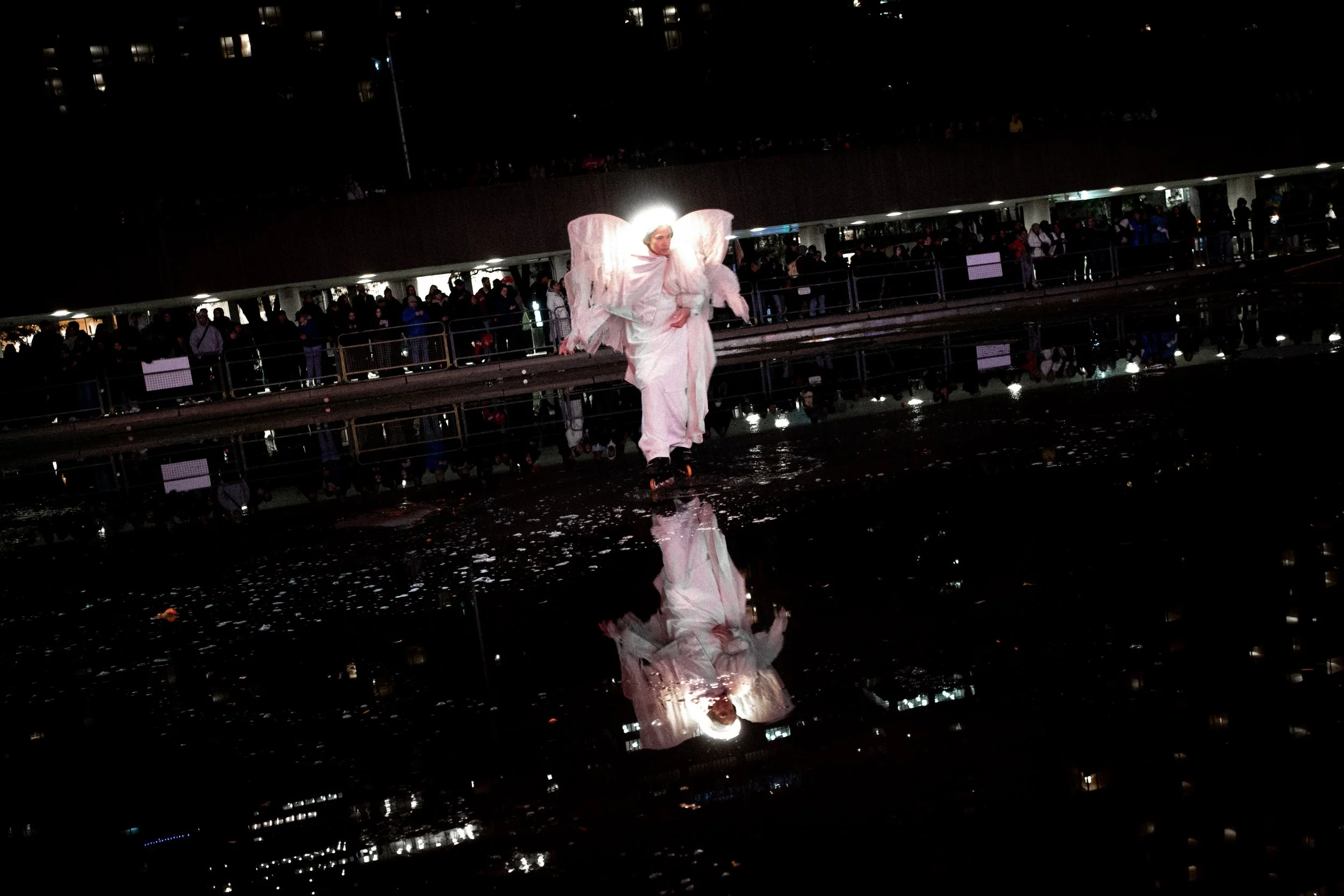 Performer dressed as an angel or fairy with large wings and halo, walking on a wet surface, with a reflective puddle showing their reflection, in front of a crowd of spectators at night.