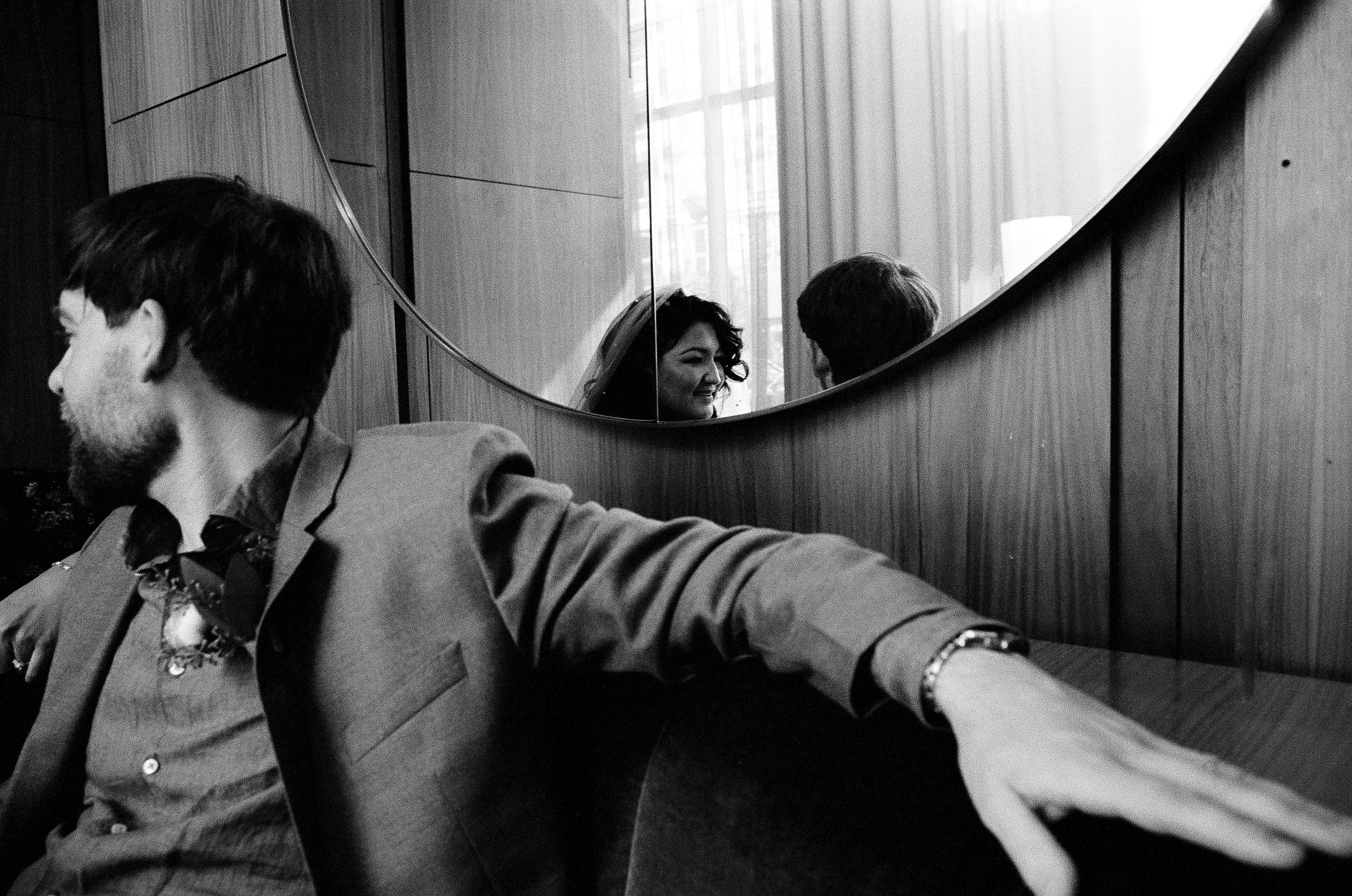 Black and white photo of three people sitting at a table, reflected in a curved mirror on a wooden wall. One woman is smiling and looking at a man with short hair. A man with glasses is on the left, looking down or away. The scene appears indoors wit