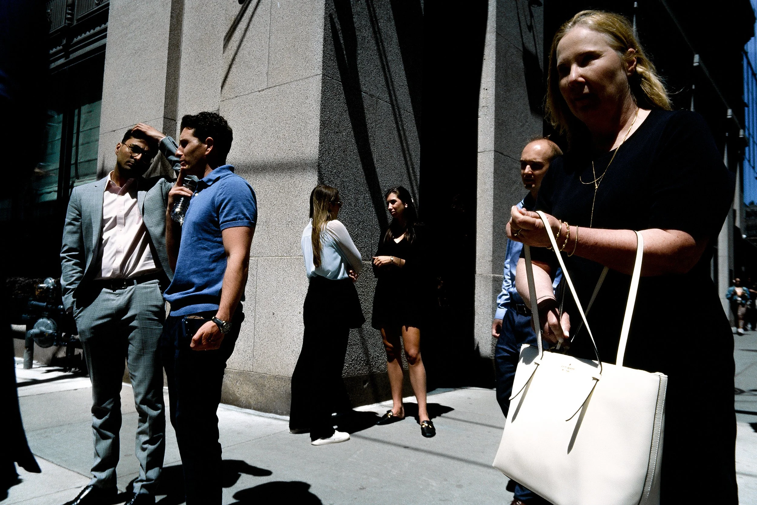 Group of people standing near a building wall, some chatting, some waiting, with shadows cast on the sidewalk.