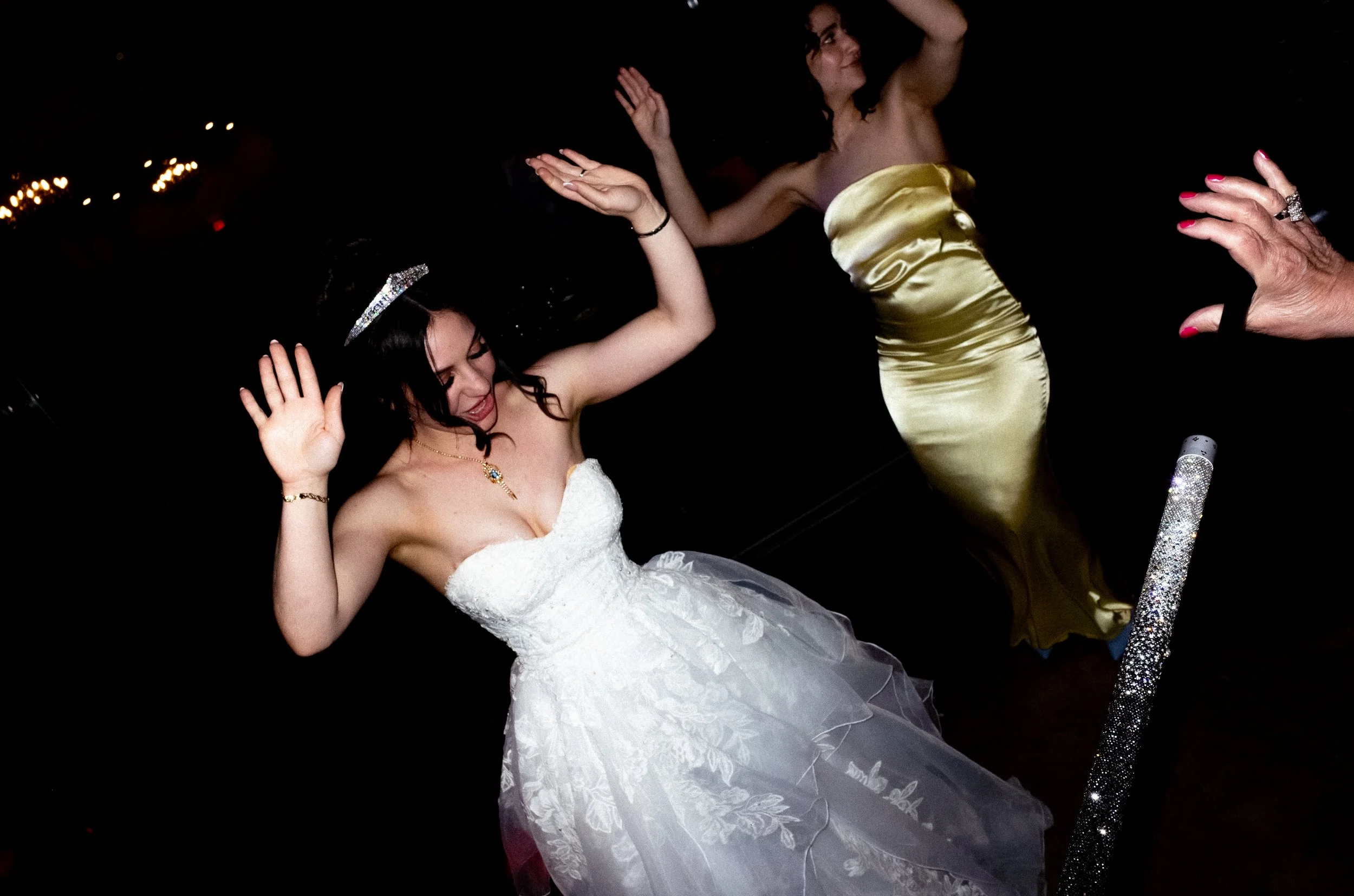 A woman in a white strapless wedding dress dancing with her eyes closed and hands raised. She is wearing a tiara, jewelry, and a gold satin dress, at a celebration or party with dark background and dim lighting.