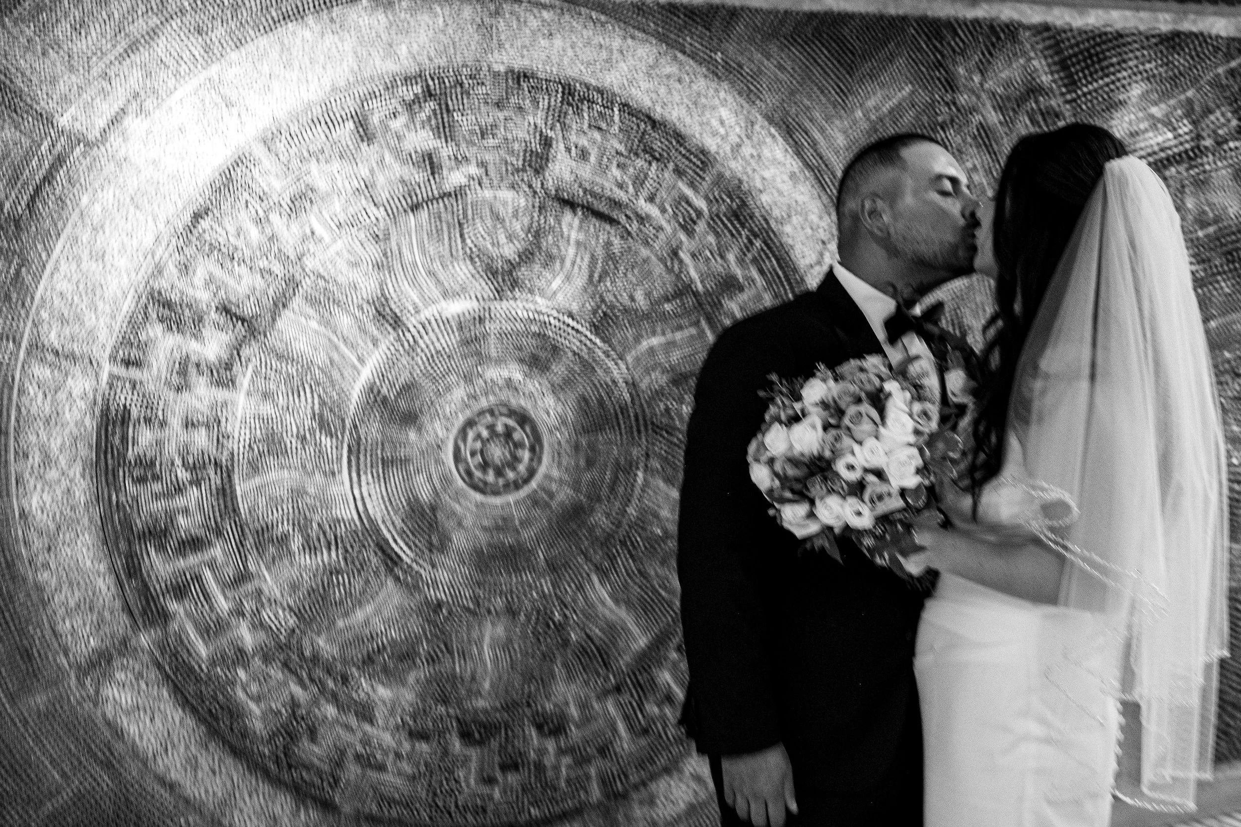 A couple in wedding attire sharing a kiss in front of a large circular textured wall with an abstract design.
