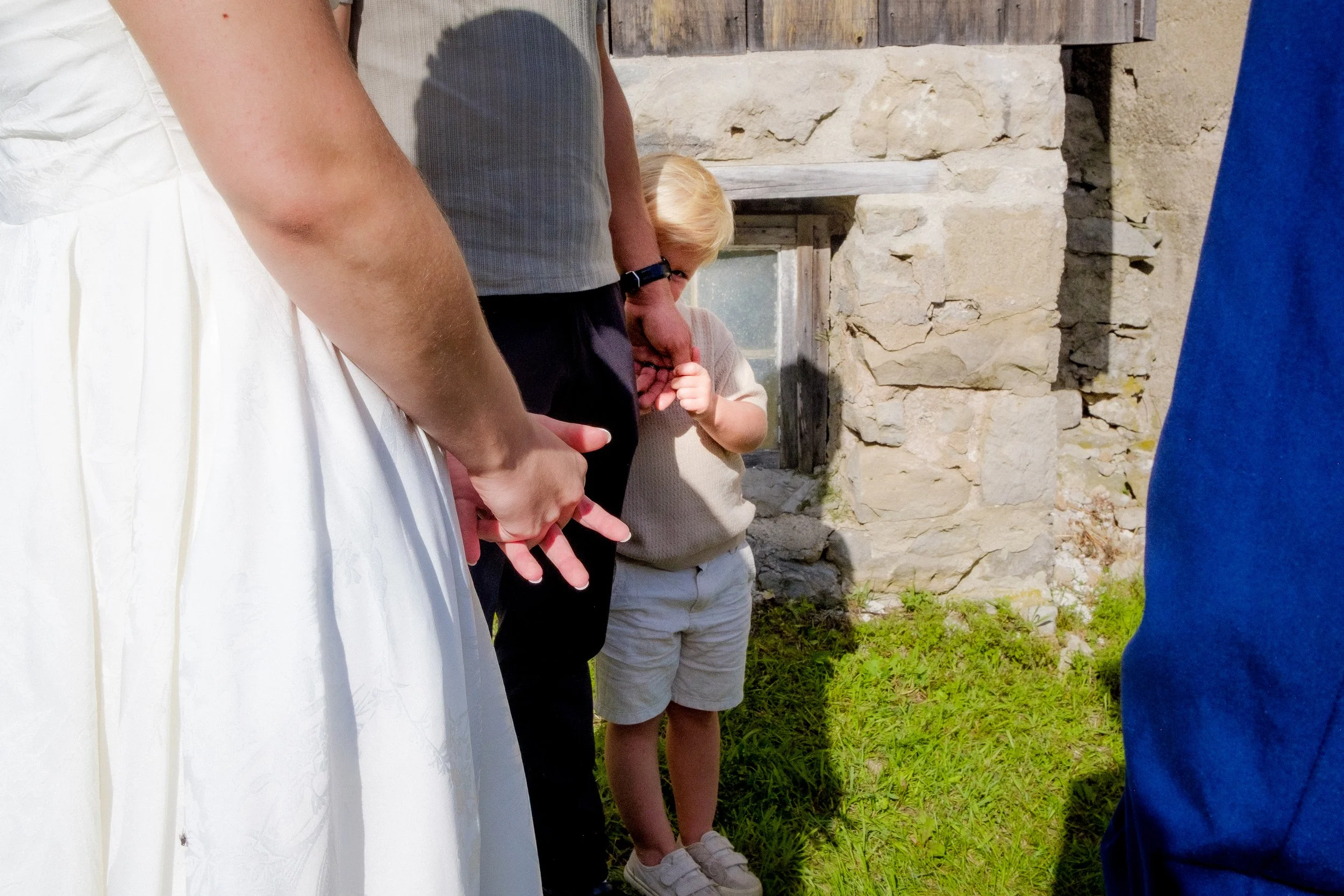 Children participating in a ceremony or outdoor event, standing close together in front of a stone wall, with one child holding their hand near their mouth.