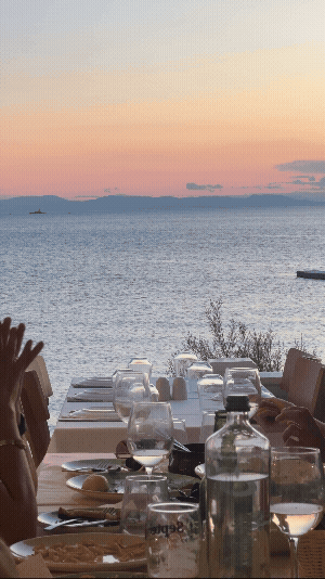Outdoor dining table set with glasses and bottles overlooking the ocean at sunset.