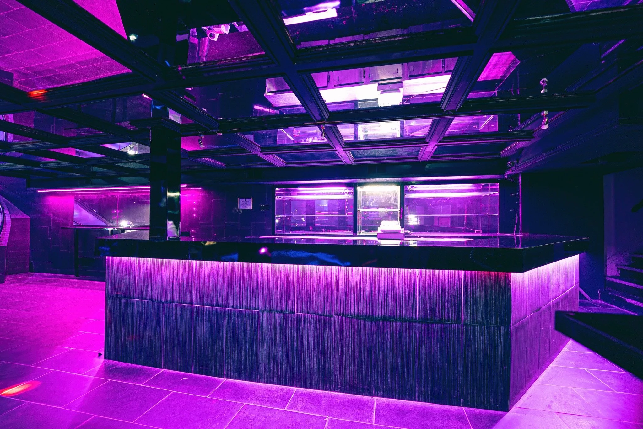 Empty nightclub bar area illuminated by purple neon lights, featuring a sleek curved bar counter with a textured black surface, tiled flooring, and reflective ceilings.