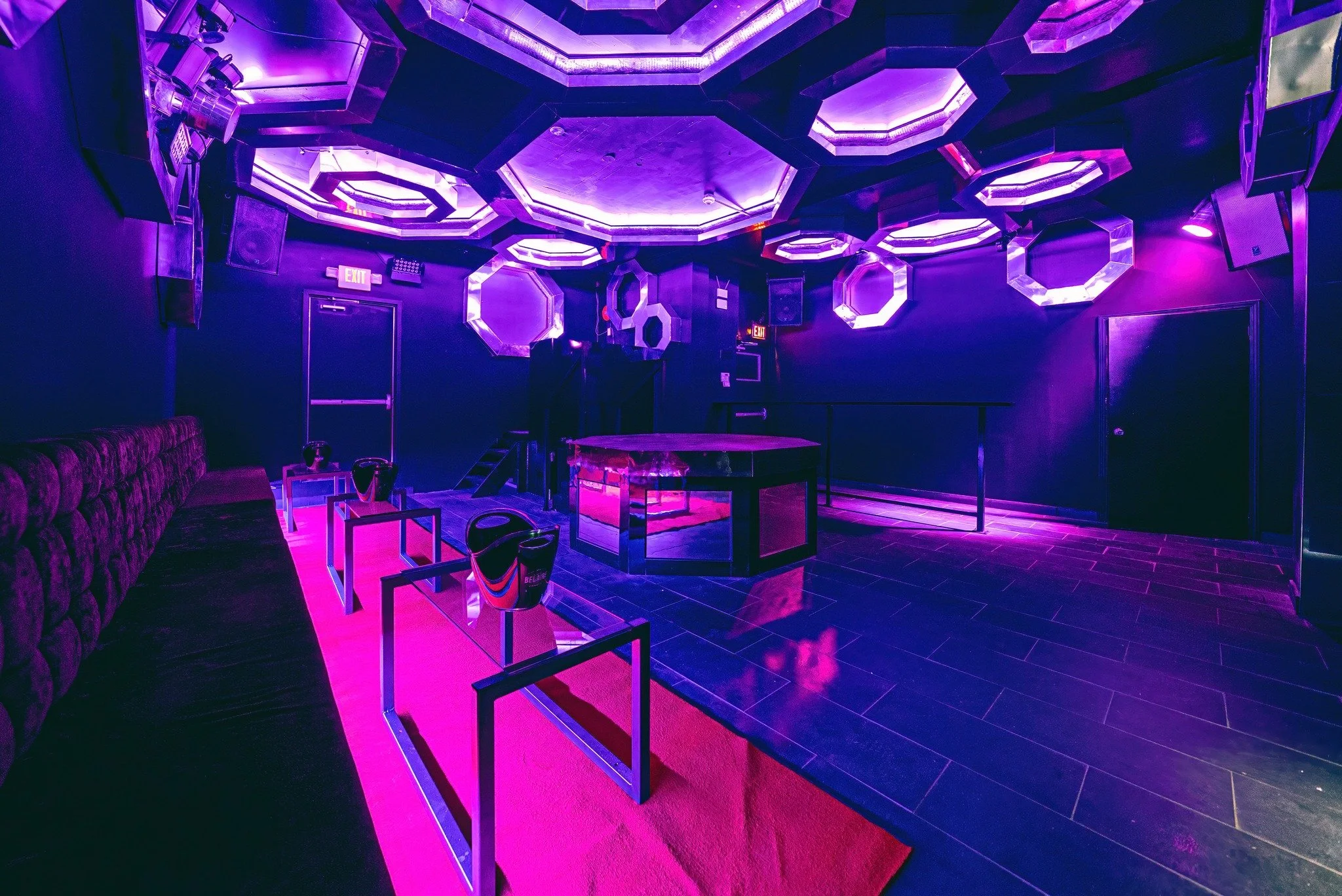 Nightclub interior with purple lighting, geometric ceiling fixtures, a dance floor, seating area with chairs and a bench, and a DJ booth or stage area.