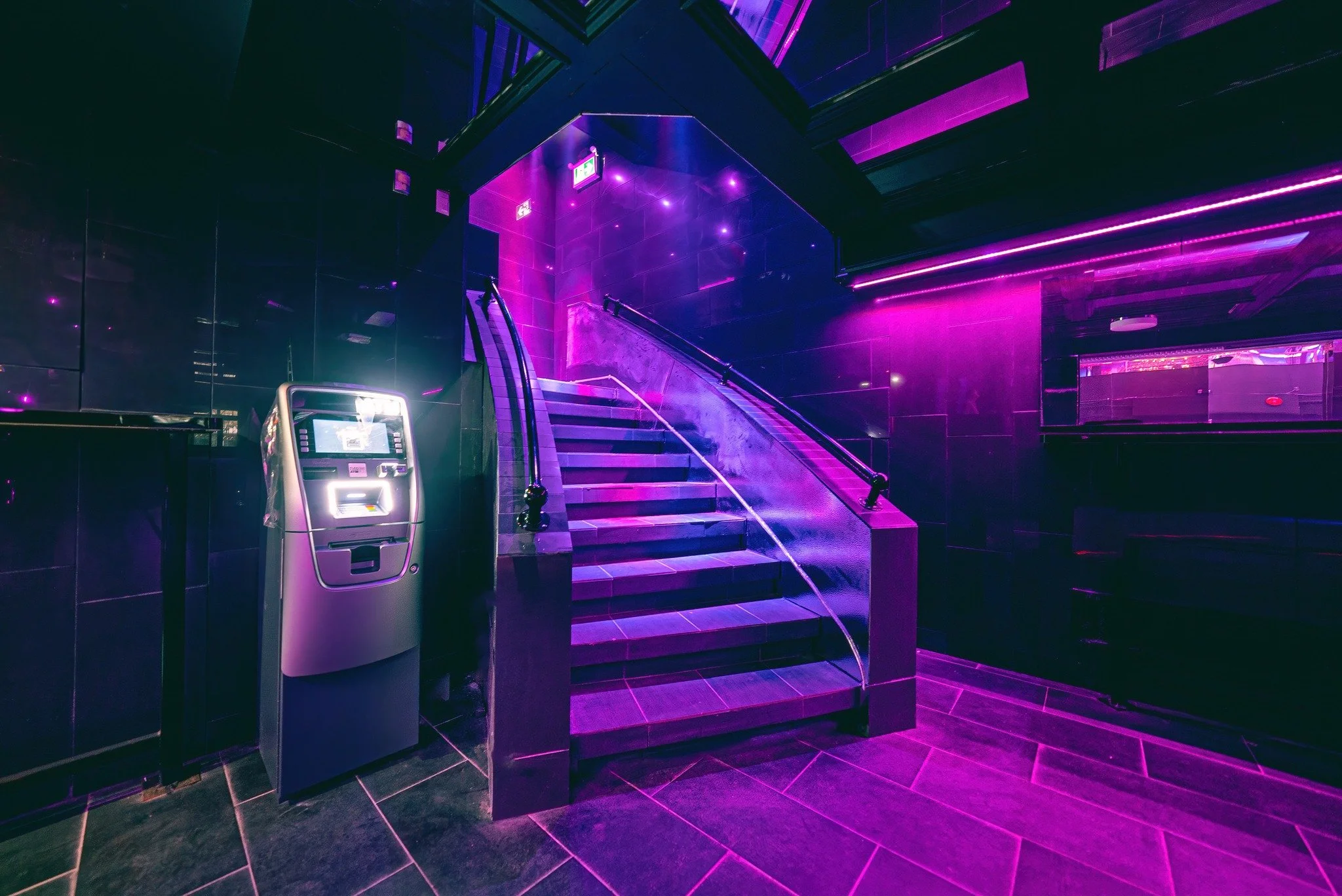 Staircase in a dimly lit space with purple and pink lighting, alongside an ATM machine.