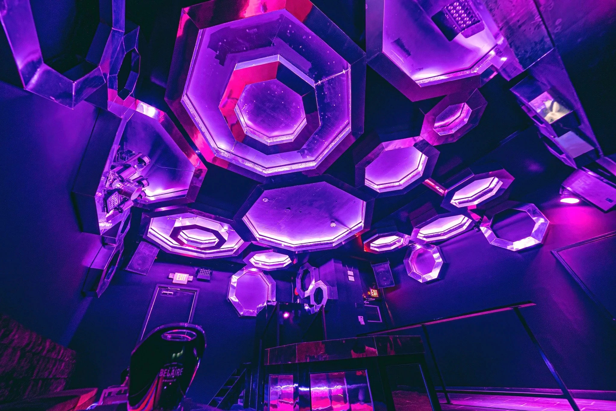 Interior of a nightclub or lounge with a purple and pink illuminated ceiling featuring hexagonal and octagonal mirrors and light fixtures.