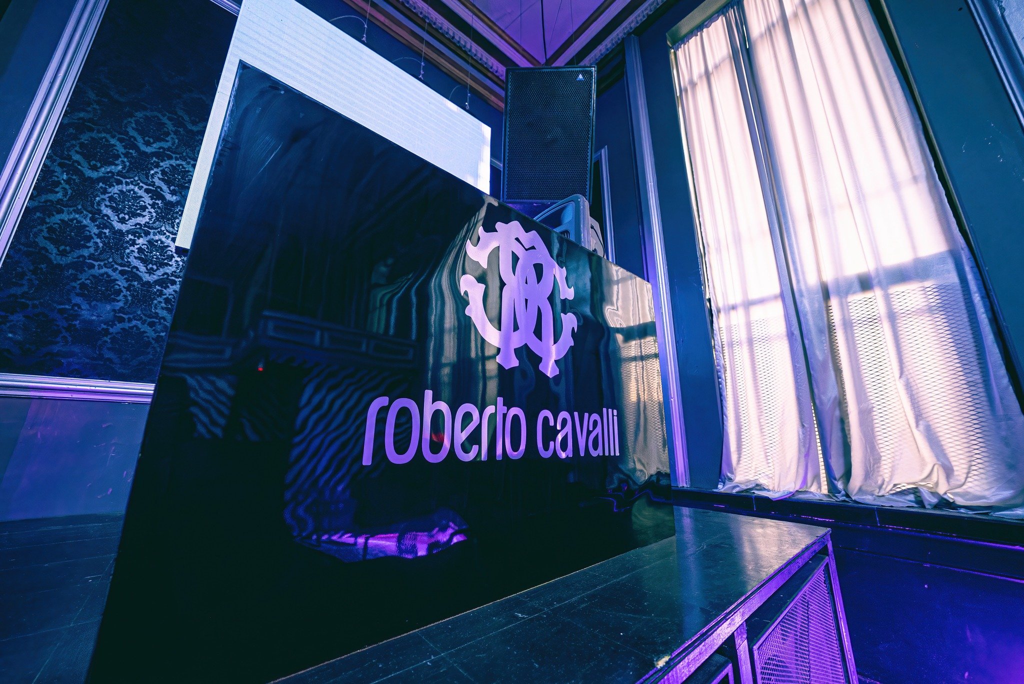 A black stage with a large sign reading 'Roberto Cavalli' and a stylized logo, set in a room with patterned and iridescent curtains and screens, under colorful lighting.