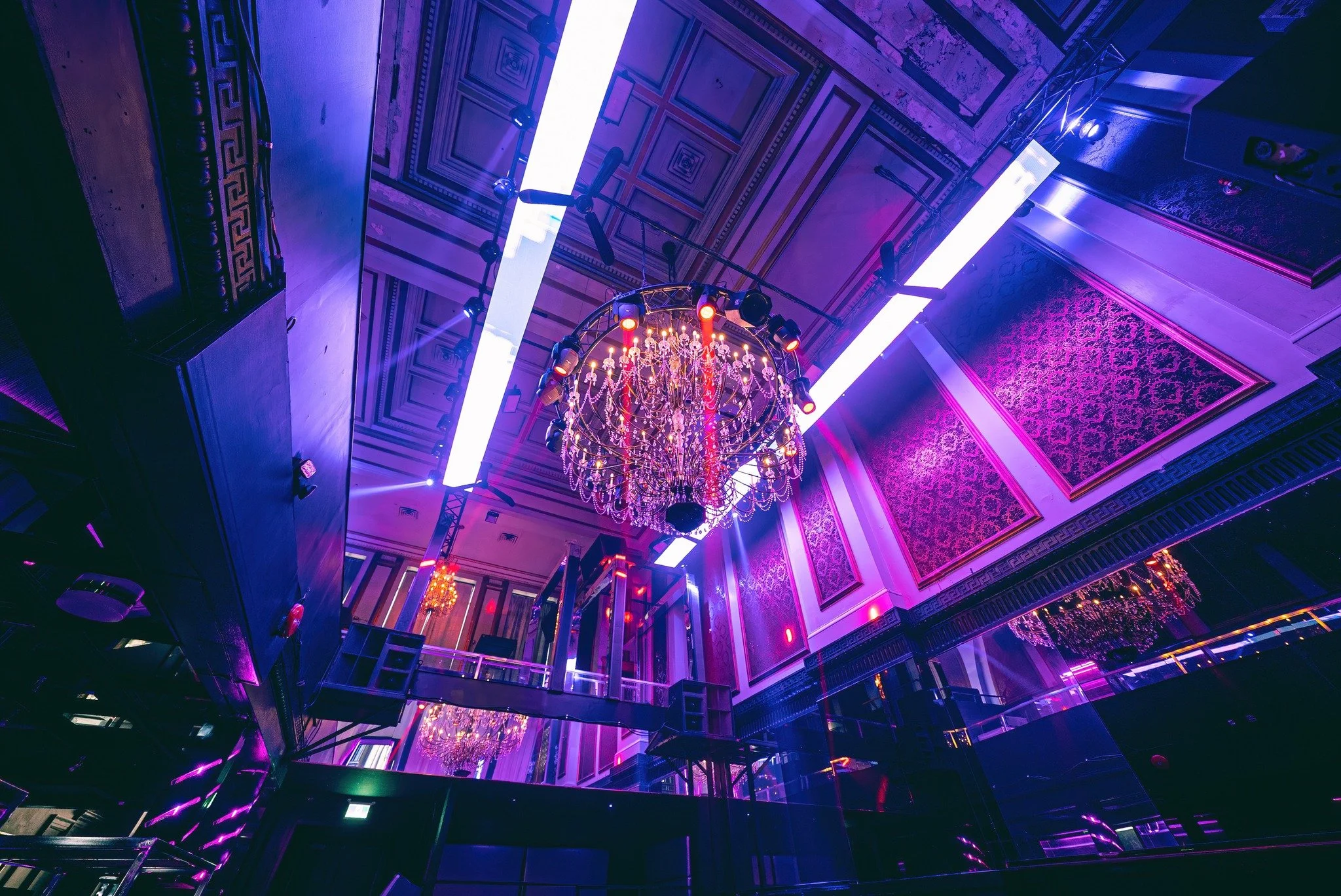 An ornate ballroom decorated with purple and pink lighting, featuring a large chandelier hanging from a high ceiling with intricate molding, and modern light fixtures.