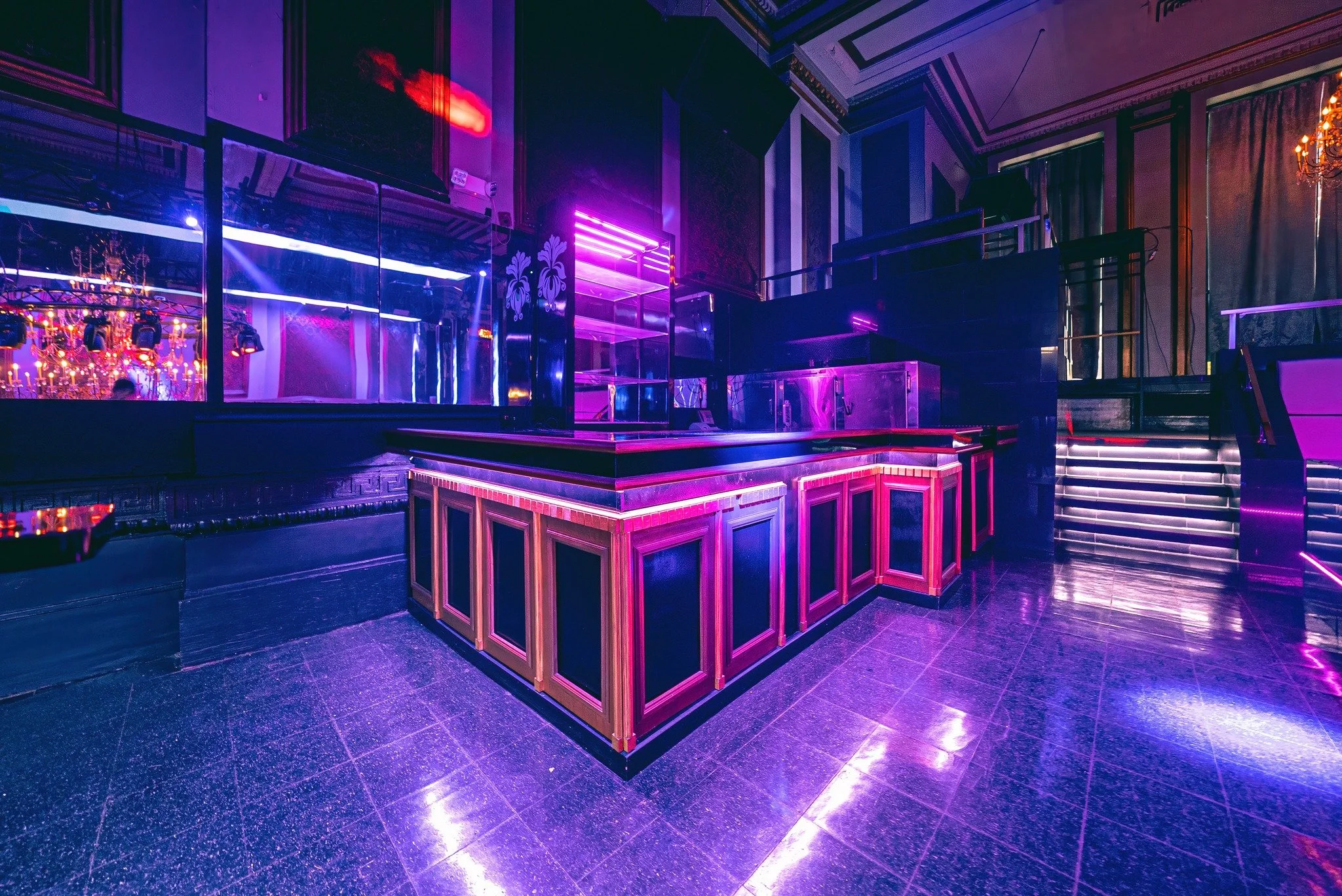 Empty nightclub bar area illuminated with purple and pink neon lights, with a curved wooden bar counter, glass display case, staircase, and chandelier.