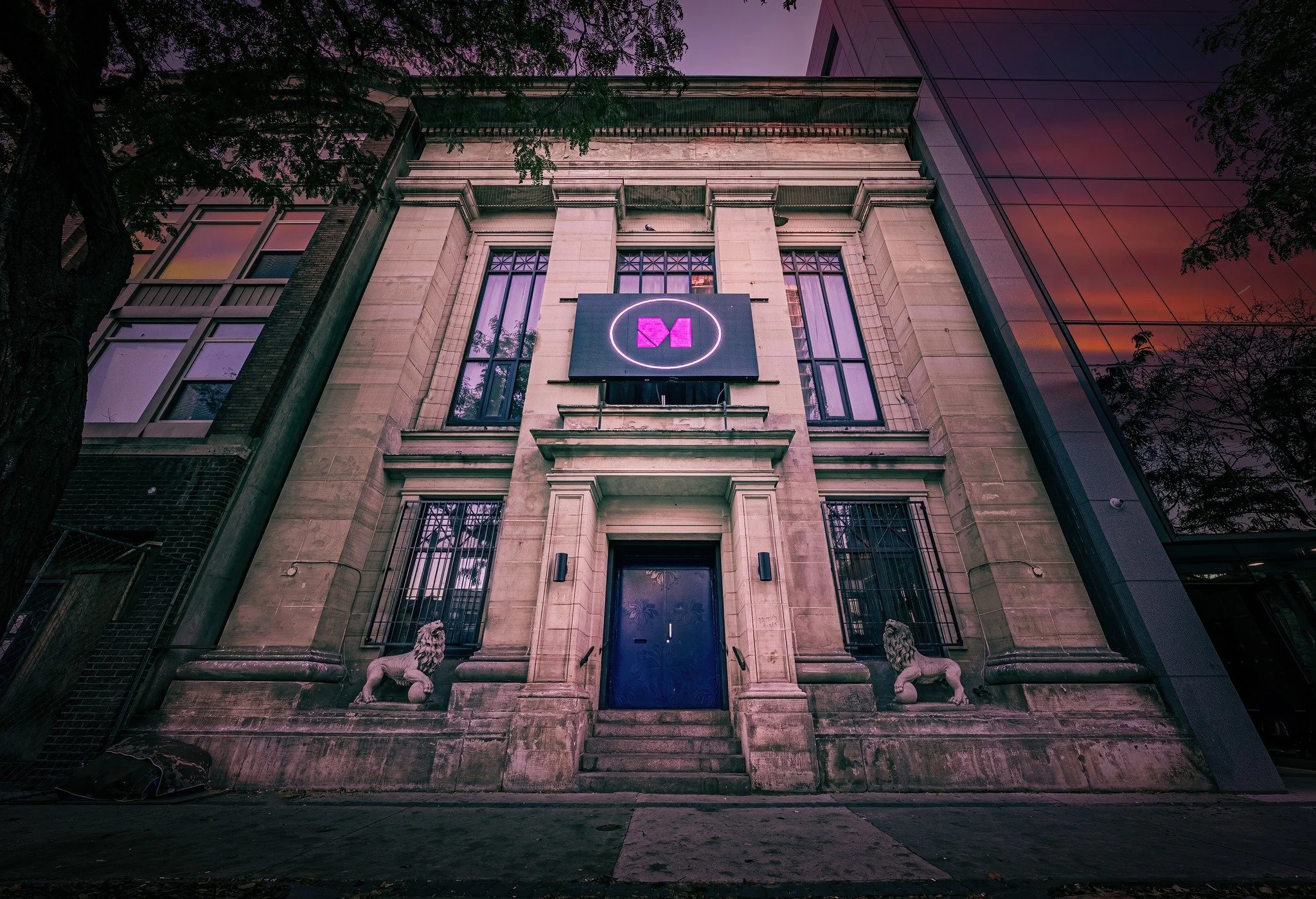 A historic brick building with modern glass extension, featuring lion statues at the entrance and a neon sign with a pink logo resembling a stylized letter 'M' above the door, during dusk.